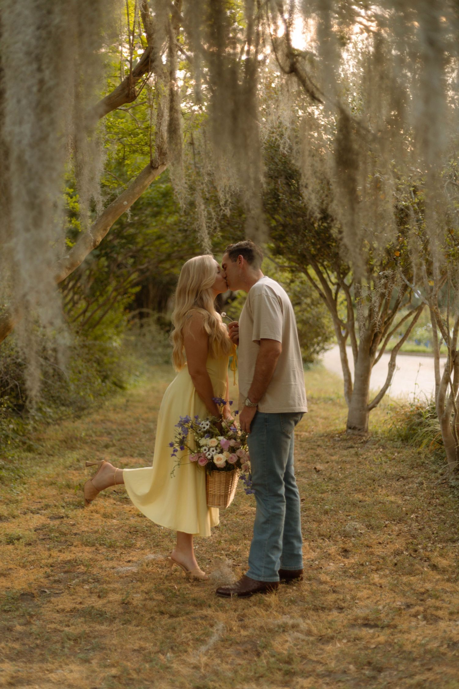 Couple kissing under Spanish moss-draped trees, woman in yellow dress holding bouquet, man in jeans, golden hour lighting.