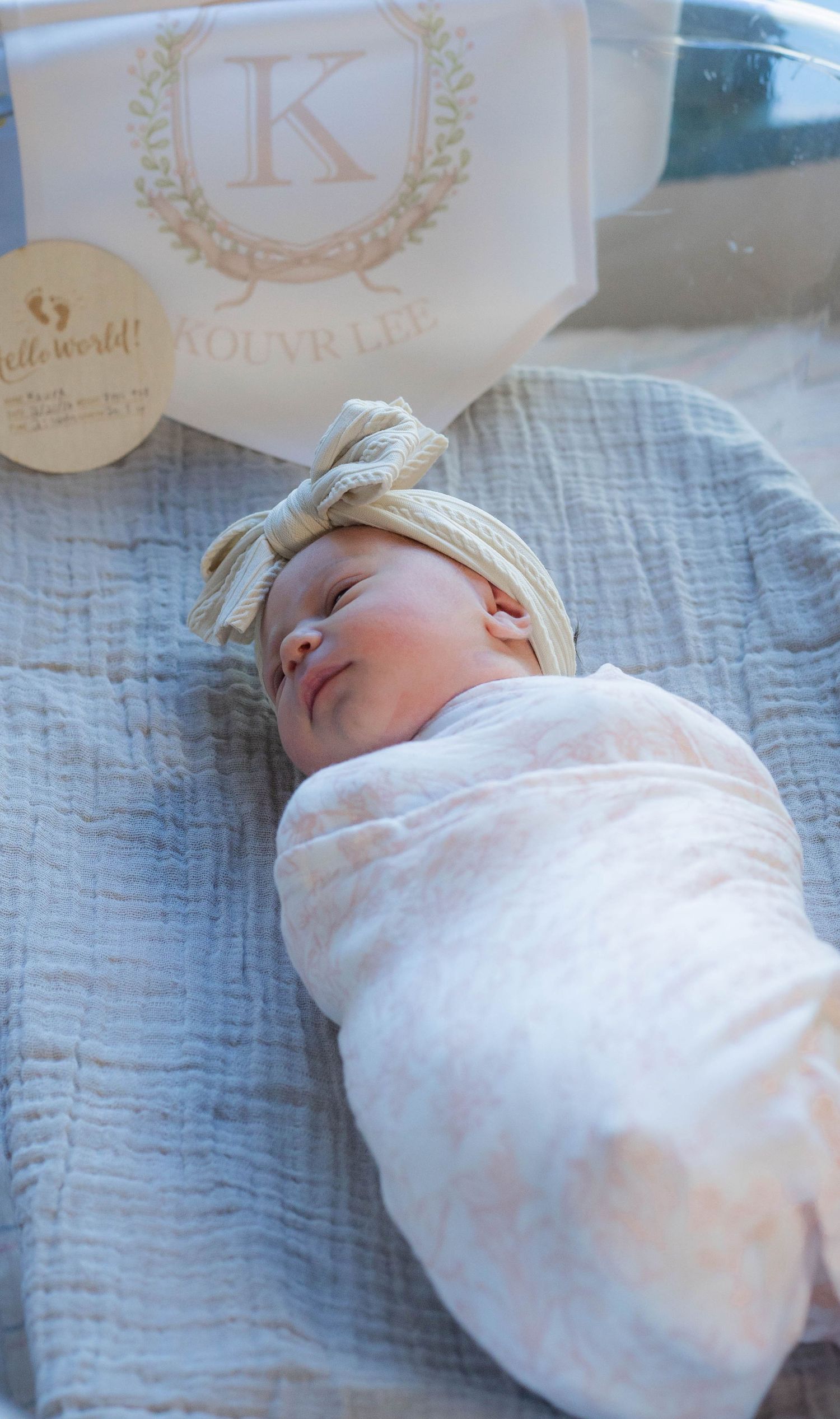 Sleeping newborn baby swaddled in white wrap with bow headband next to a personalized name sign reading Kolyr Lee.