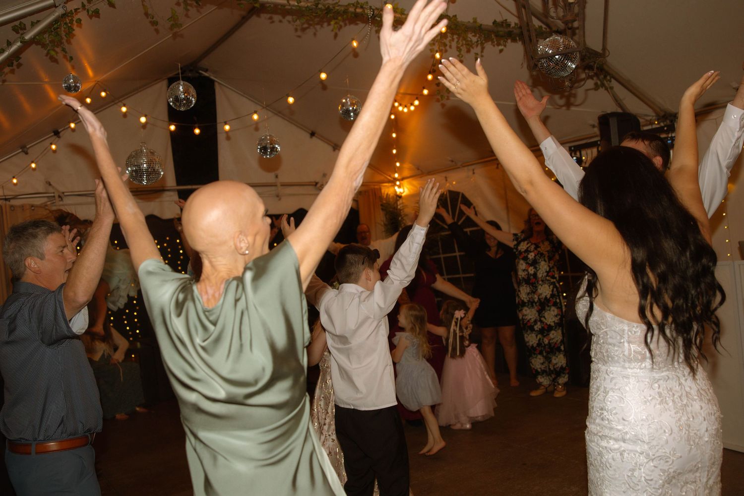 Guests dancing joyfully at a wedding reception under string lights and a disco ball in a decorated tent.