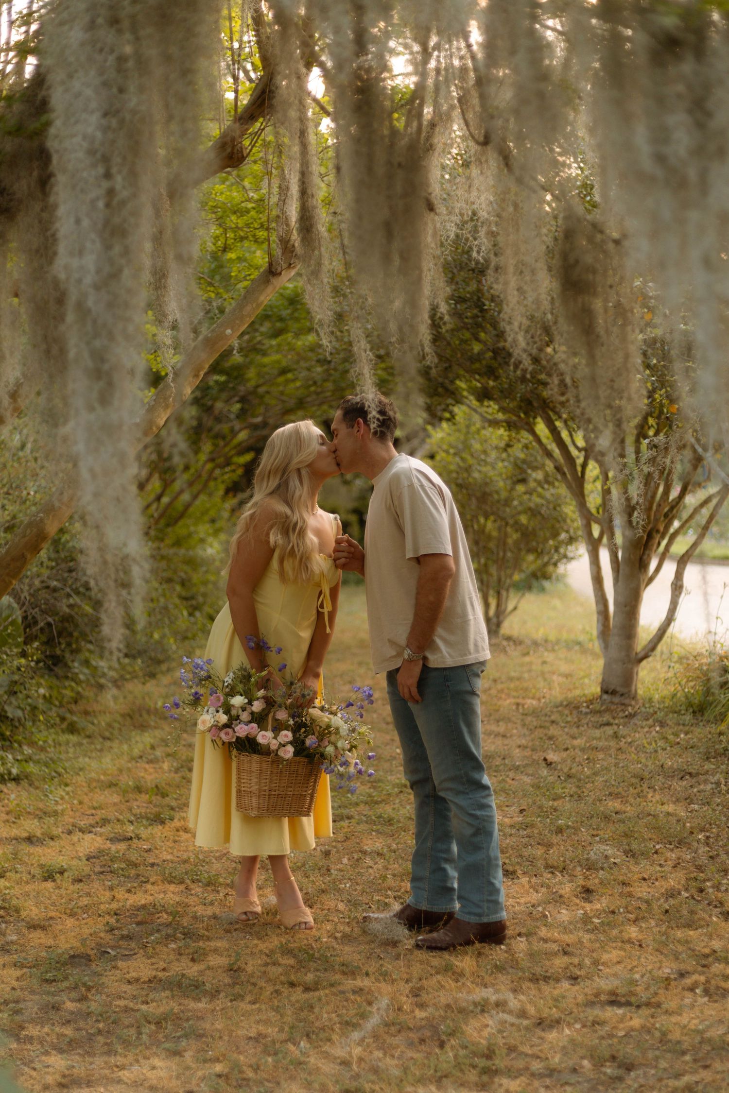 A couple shares a romantic kiss under Spanish moss-draped trees, she holds a wildflower basket in a yellow dress.