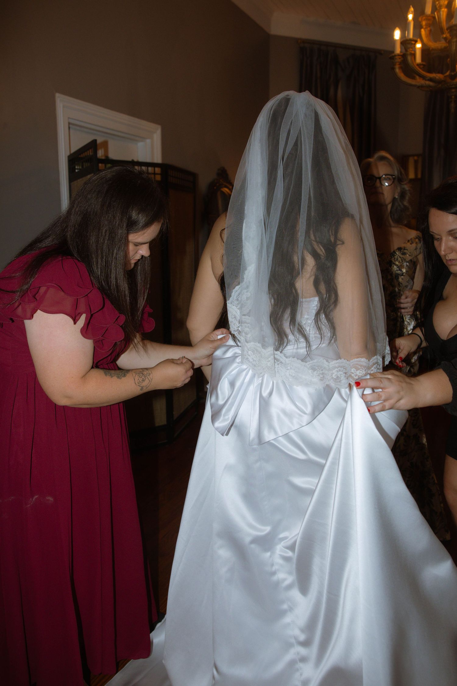 Bridesmaids helping bride with white satin wedding dress and veil before ceremony in elegant room.