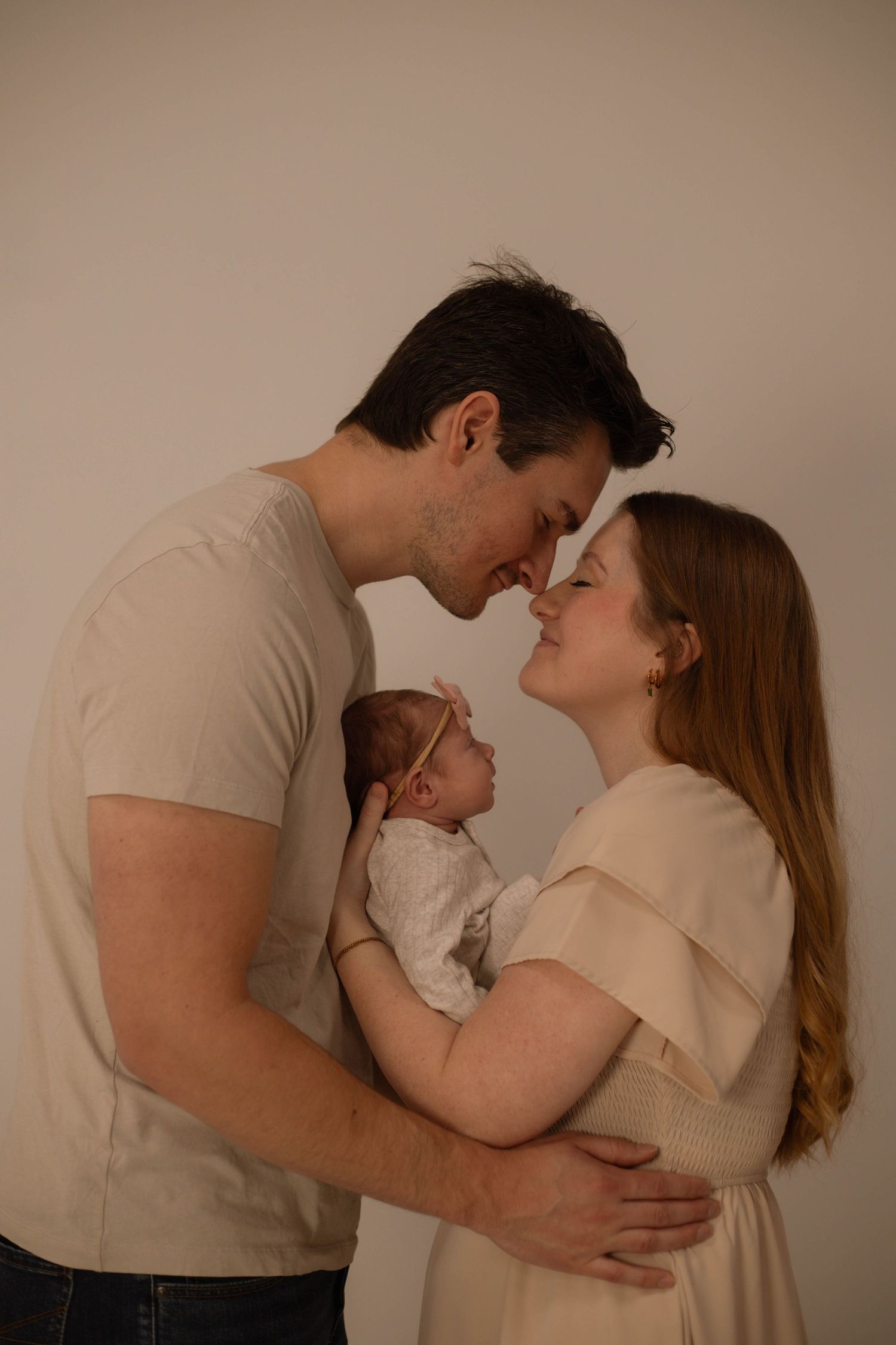 A couple shares a tender nose-to-nose moment while holding their newborn baby, dressed in neutral tones against a soft background.