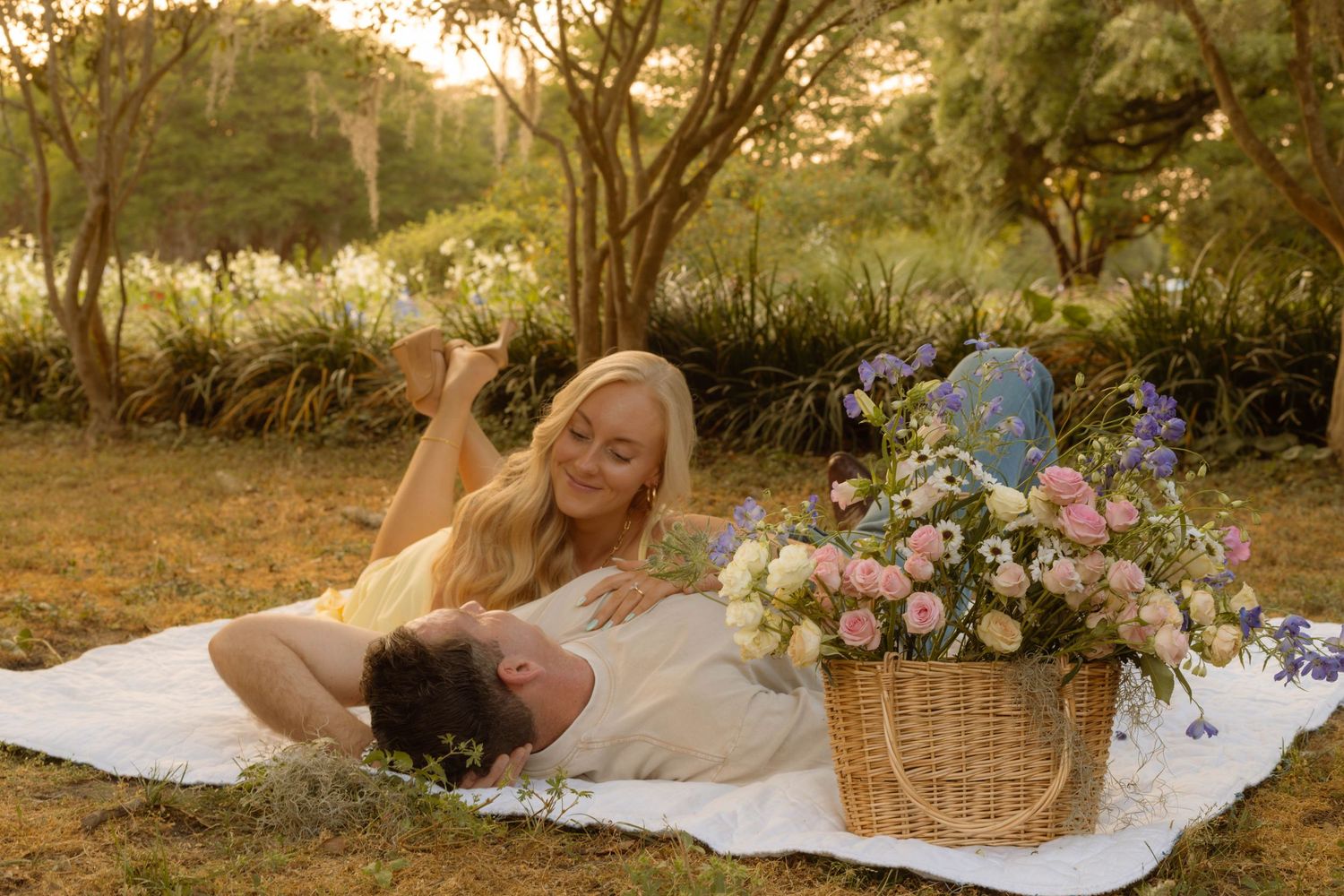 Couple relaxing on white blanket in golden hour park setting with floral basket nearby.