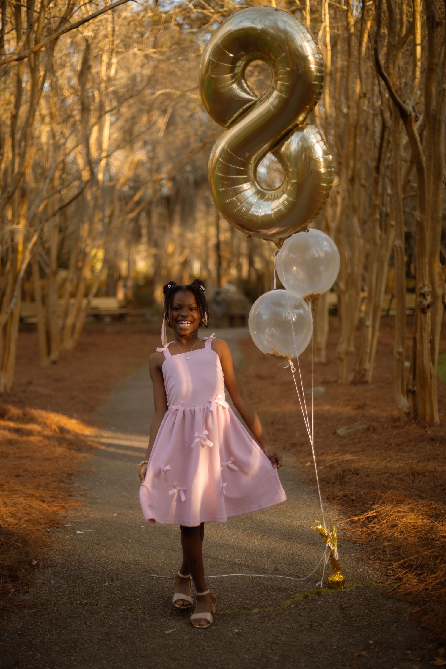 A smiling girl in a pink dress holds balloons including a gold number 8 balloon on a tree-lined path.