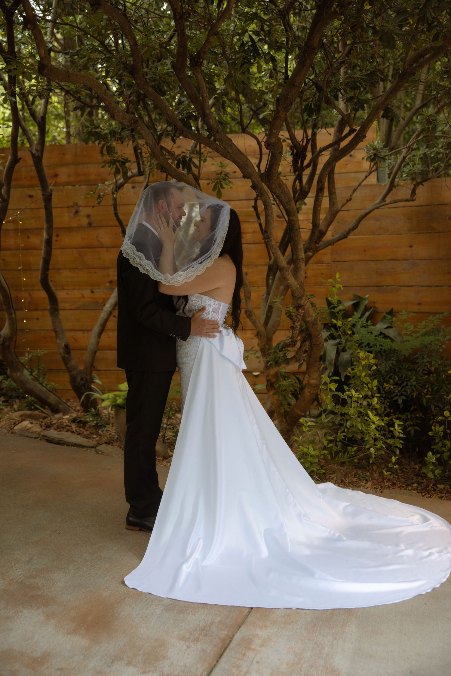 Bride and groom share a kiss under a veil in a garden with wooden fence backdrop.