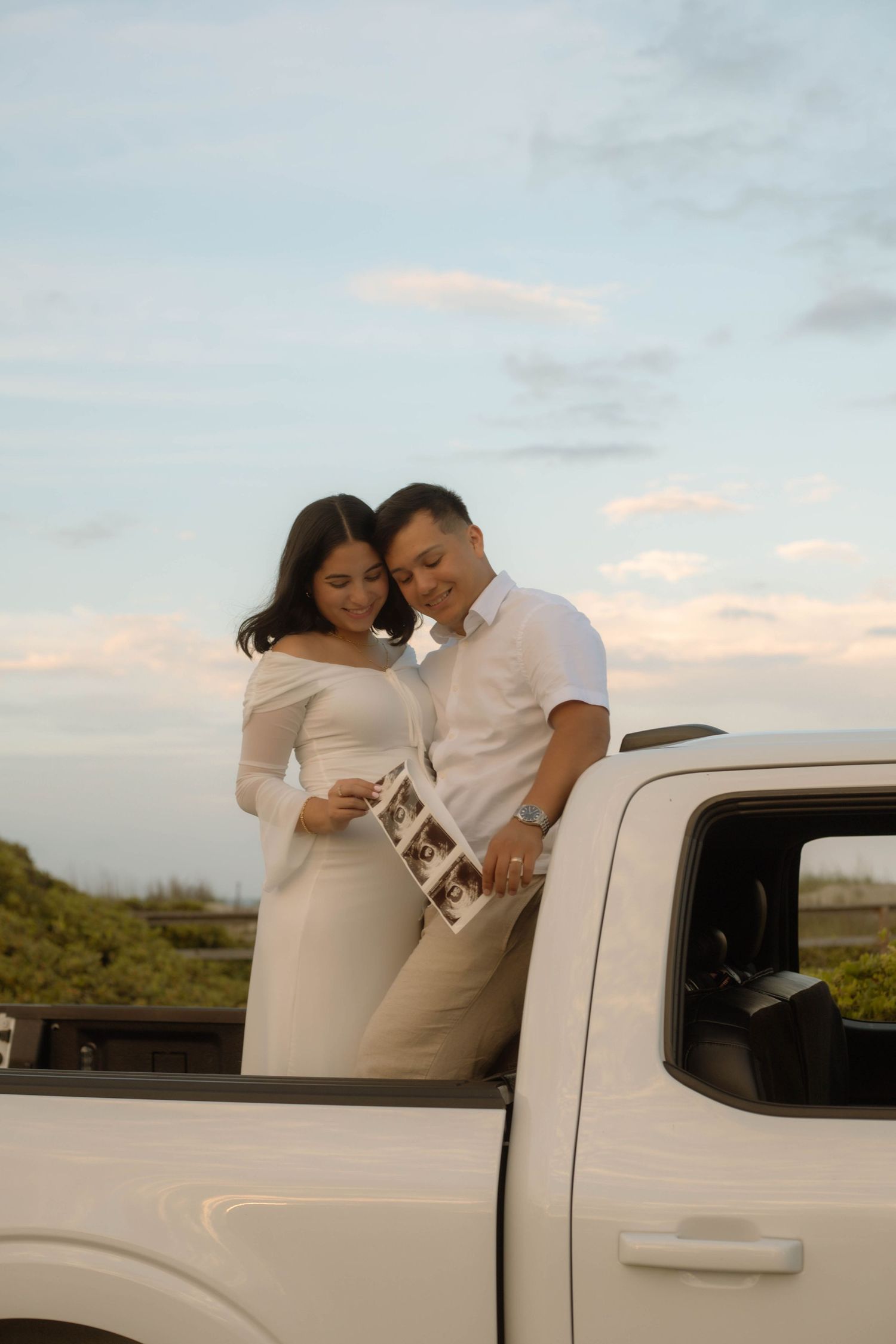 Expecting couple in white outfits holding ultrasound photos beside a white pickup truck at sunset.