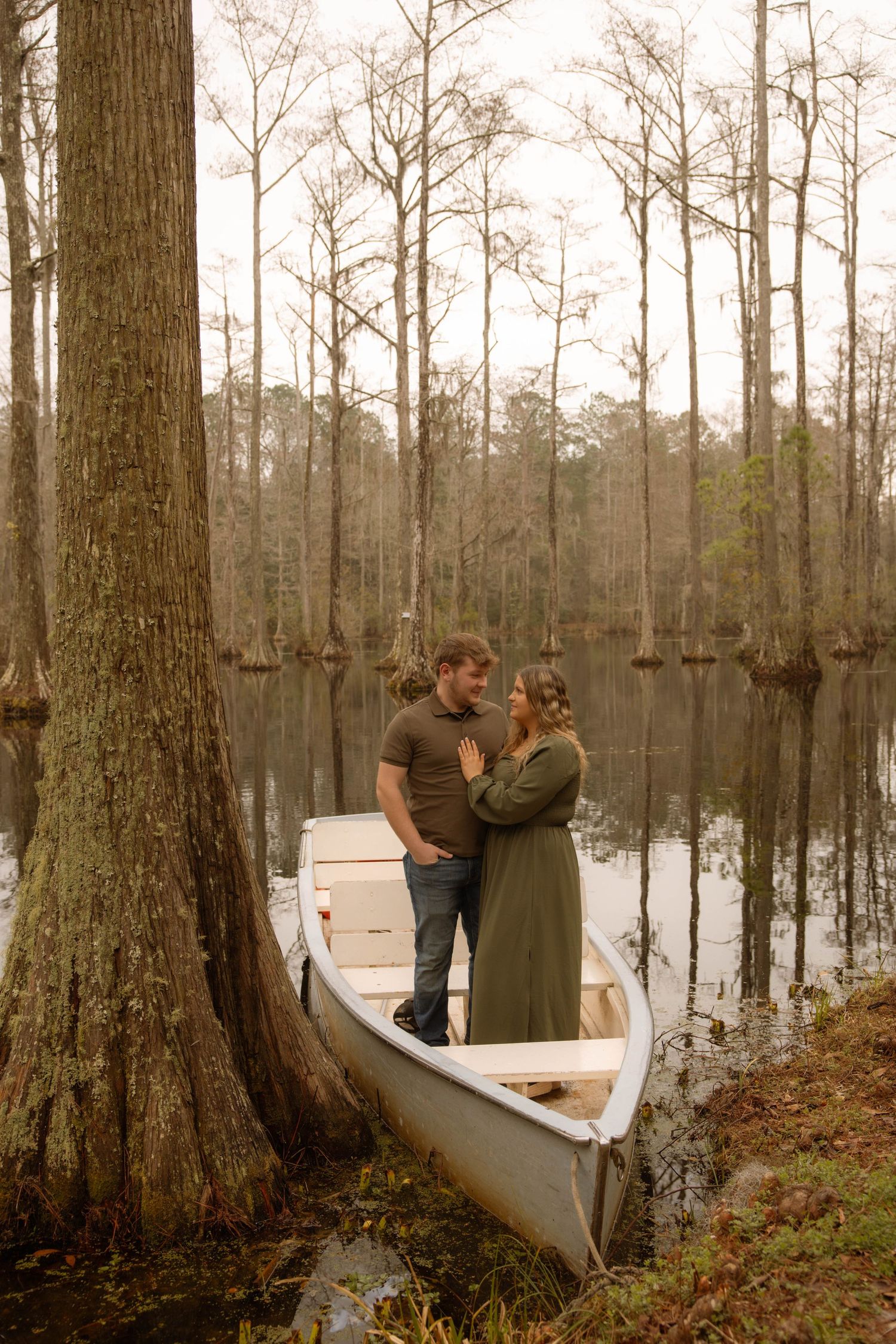 Couple standing romantically in a canoe by a cypress swamp, surrounded by tall bare trees reflecting in still water.