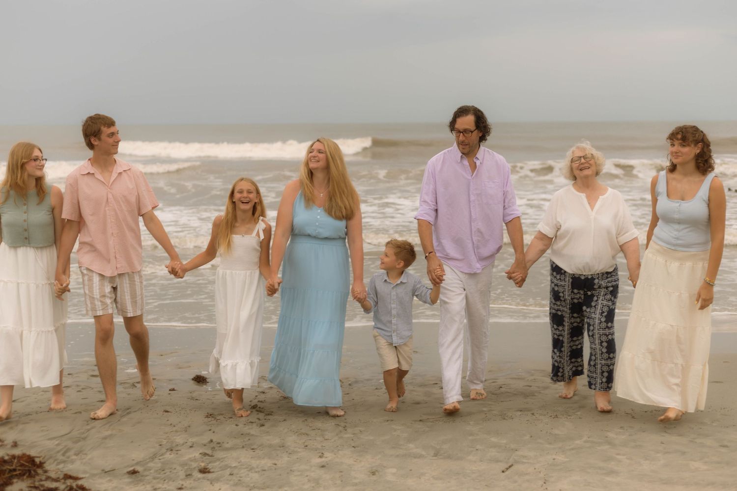 Multi-generational family holding hands walking along a sandy beach with ocean waves in the background.