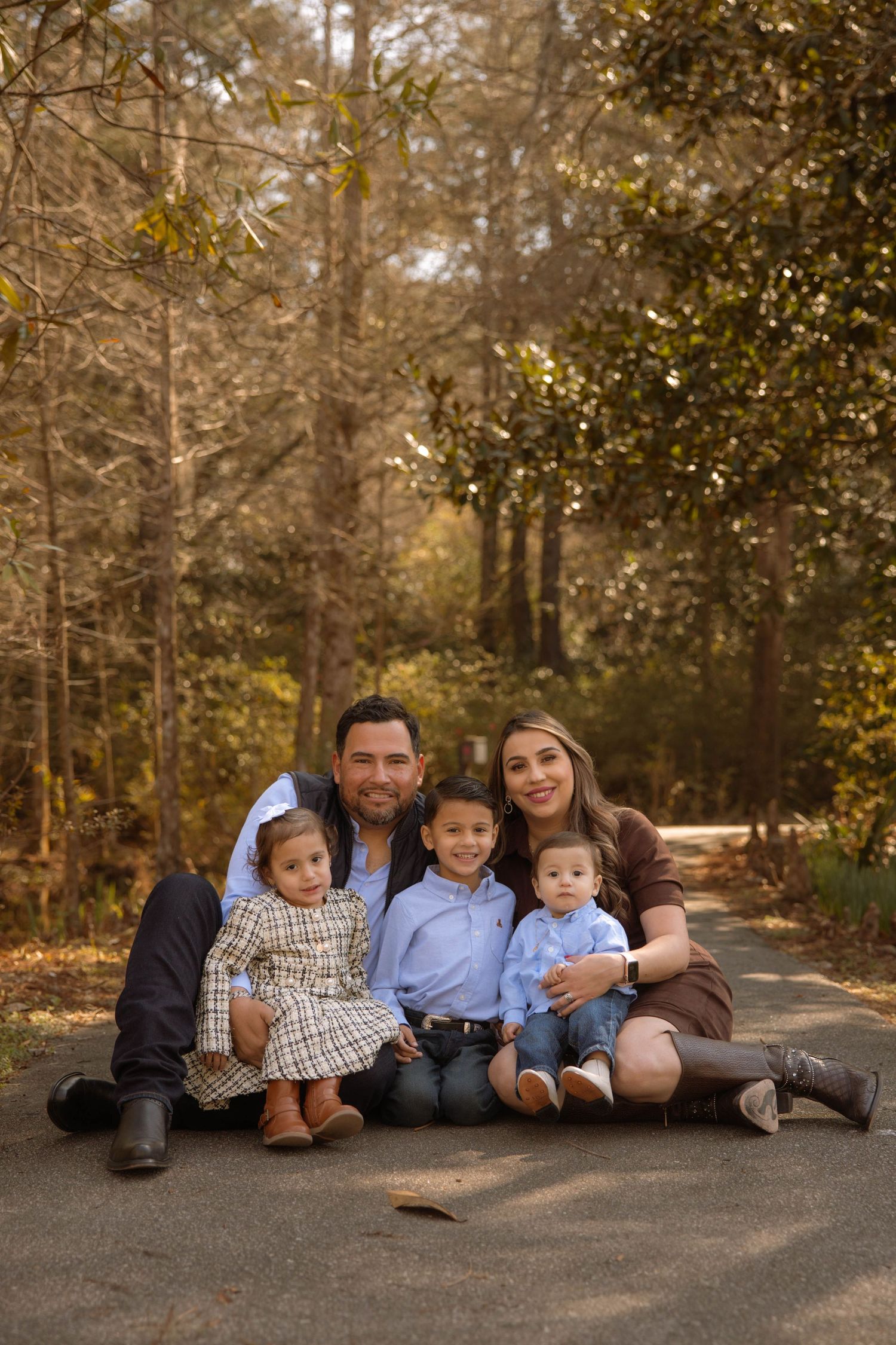 Family of five smiling together outdoors on a path surrounded by autumn trees in a warm, natural setting.