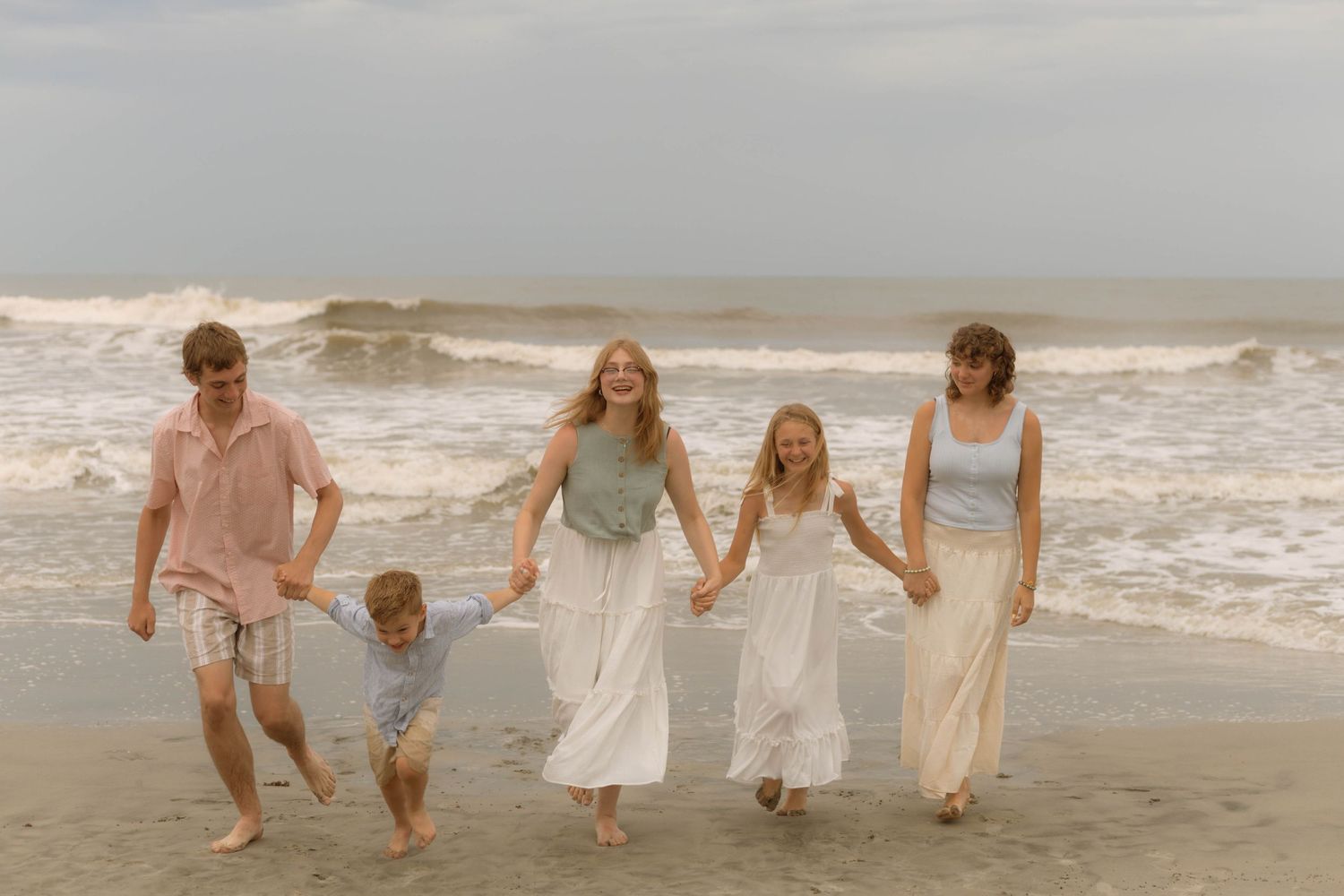Family of five walking hand-in-hand on a beach, waves crashing behind them on a cloudy day.