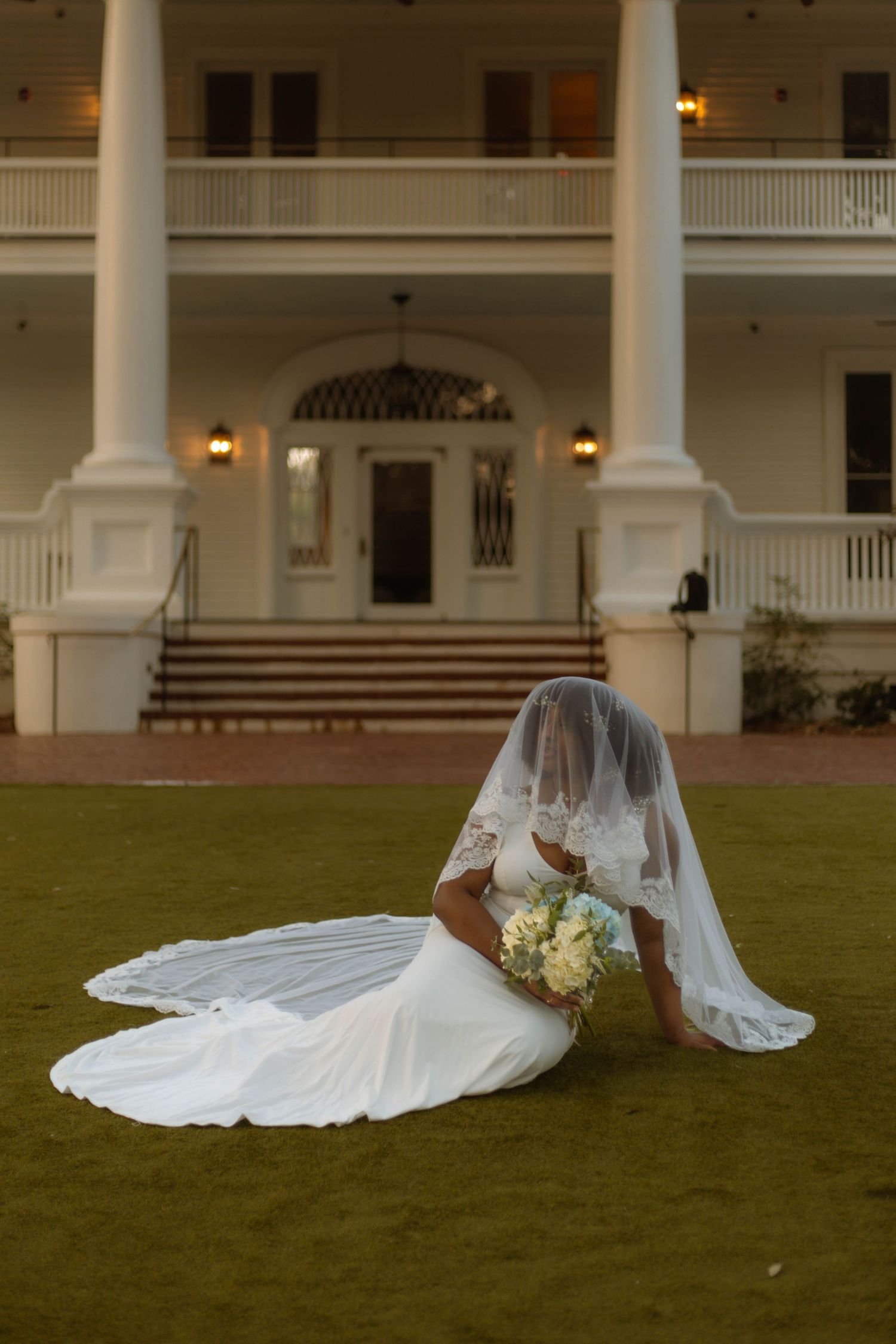 A long white wedding dress train with delicate lace details spreads across a manicured lawn in front of a colonial building.