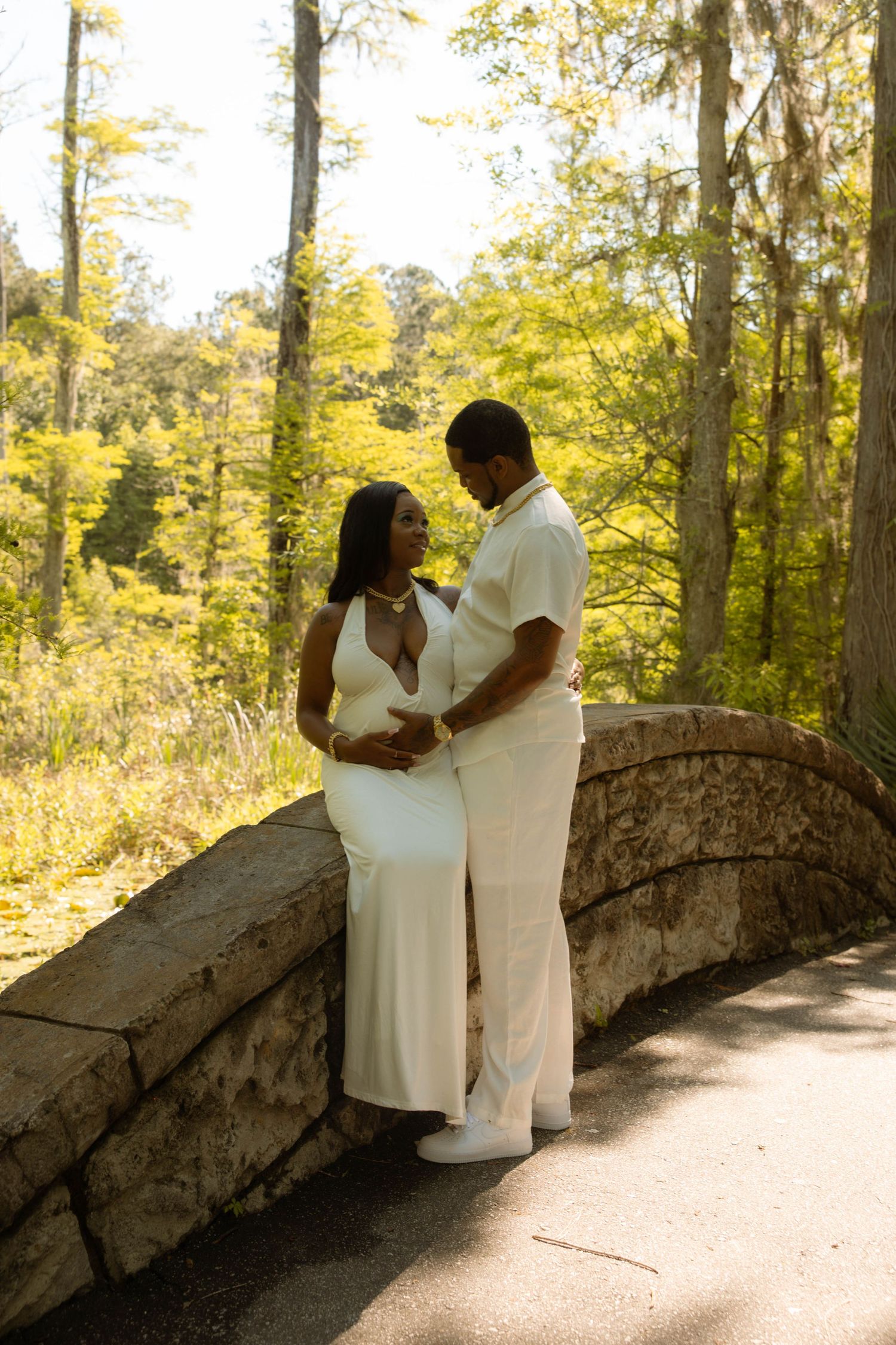 Pregnant couple in matching white outfits embracing on a stone bridge surrounded by lush green trees in a park.