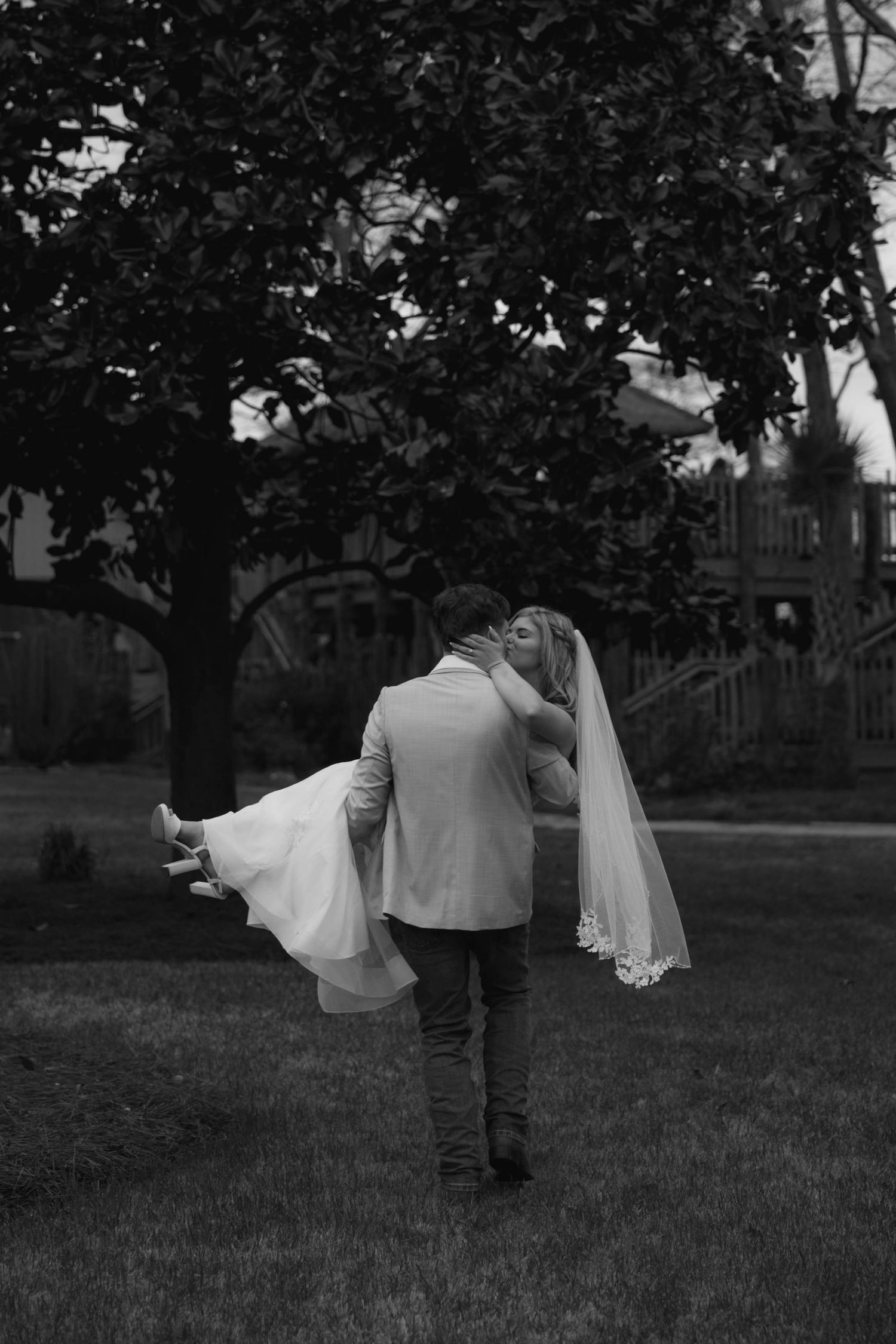 Groom carries bride in wedding dress and veil while sharing a kiss outdoors in black and white photo.