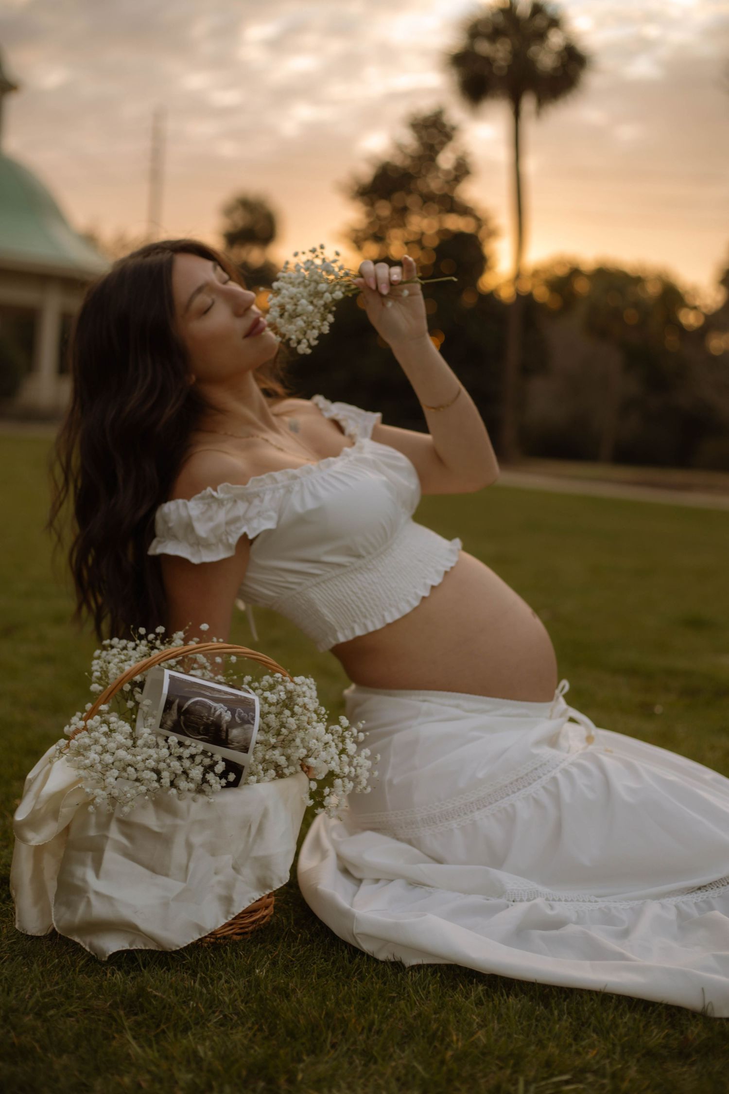 Pregnant woman in white crop top and skirt sits on grass at sunset, holding baby's breath flowers beside a basket with ultrasound photos.
