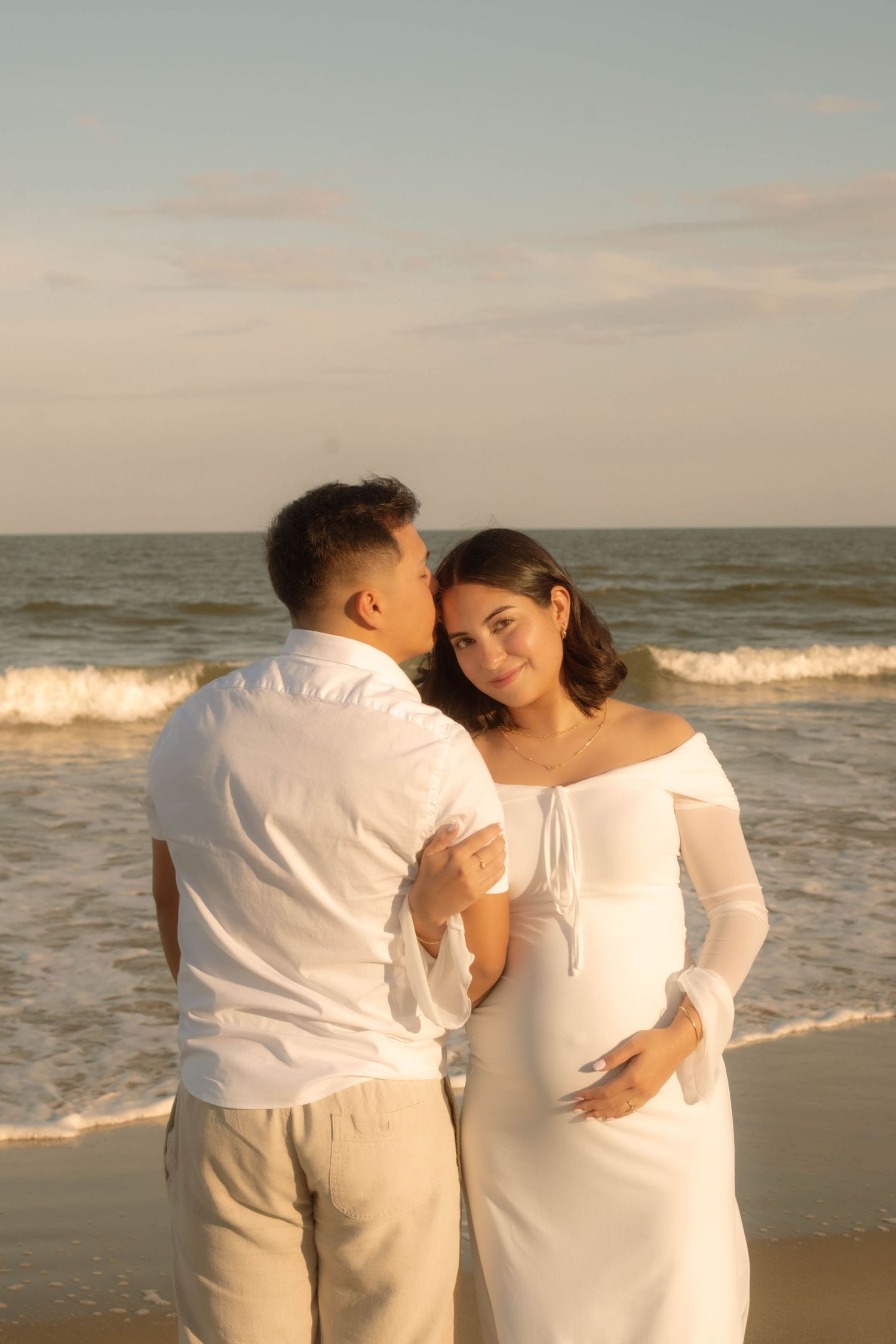 Pregnant couple in white outfits sharing a tender moment on a beach at golden hour, waves in background.