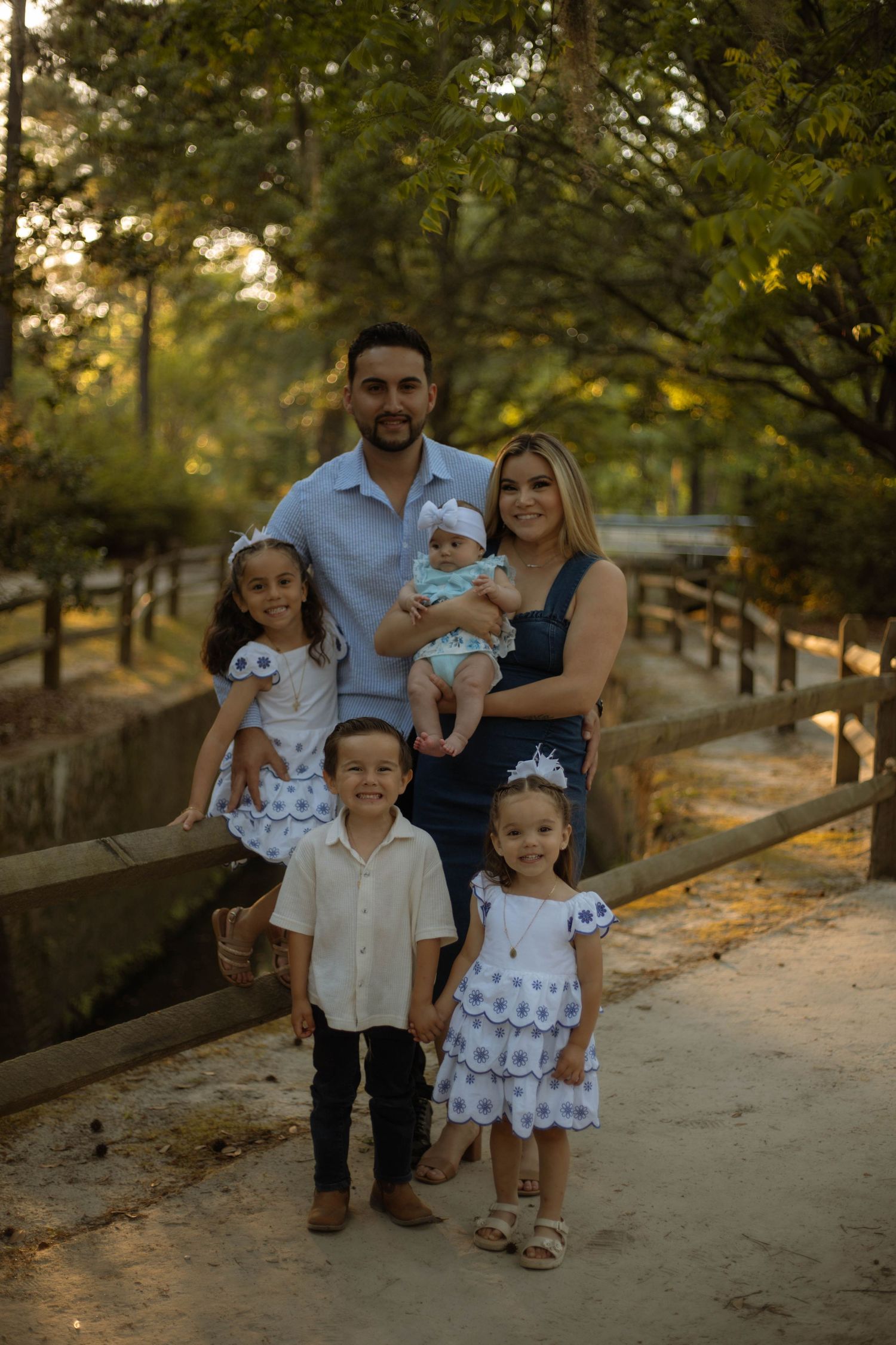 Family of five posing outdoors on a sunny path surrounded by trees, parents with three young children dressed in white.