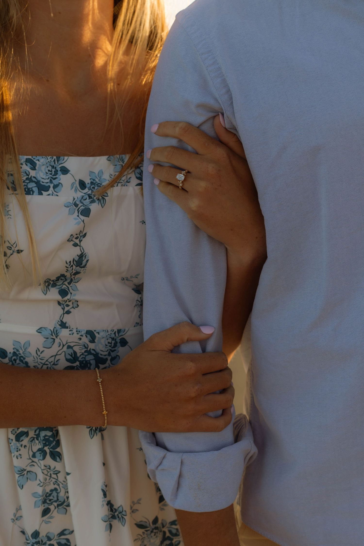 Woman in floral dress wearing an engagement ring holds man's arm in warm golden light.