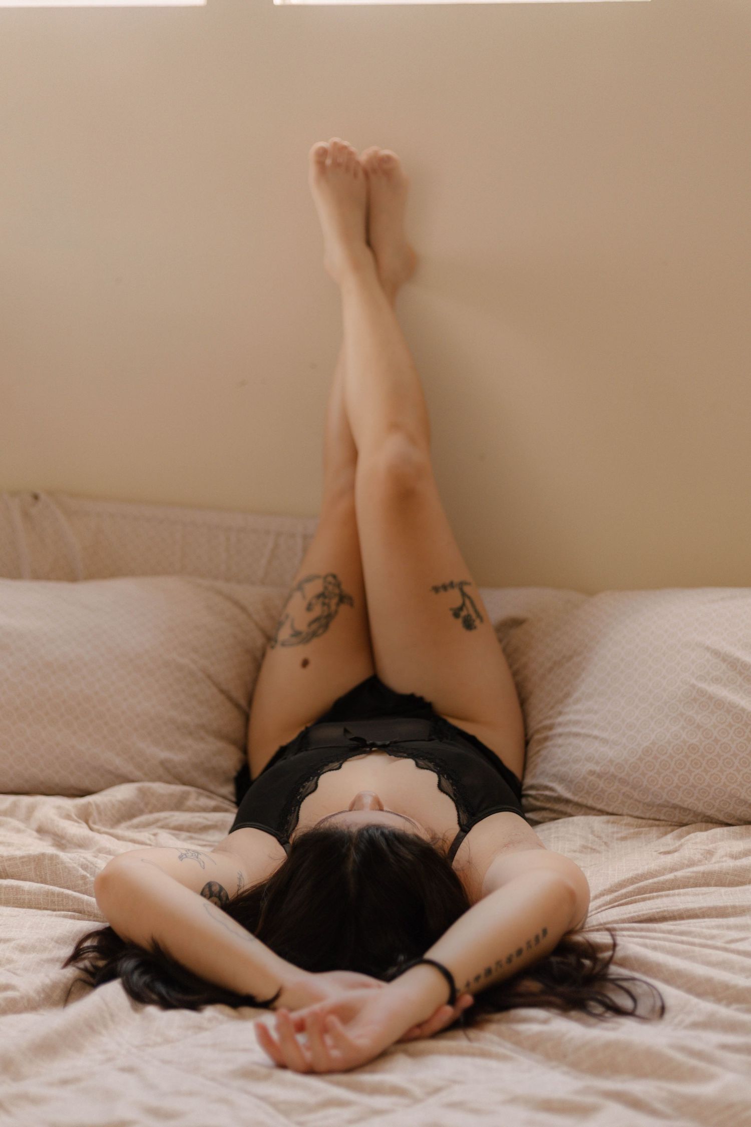 Woman in black lingerie lying on bed with legs raised against wall, showing tattoos on thigh and wrist.