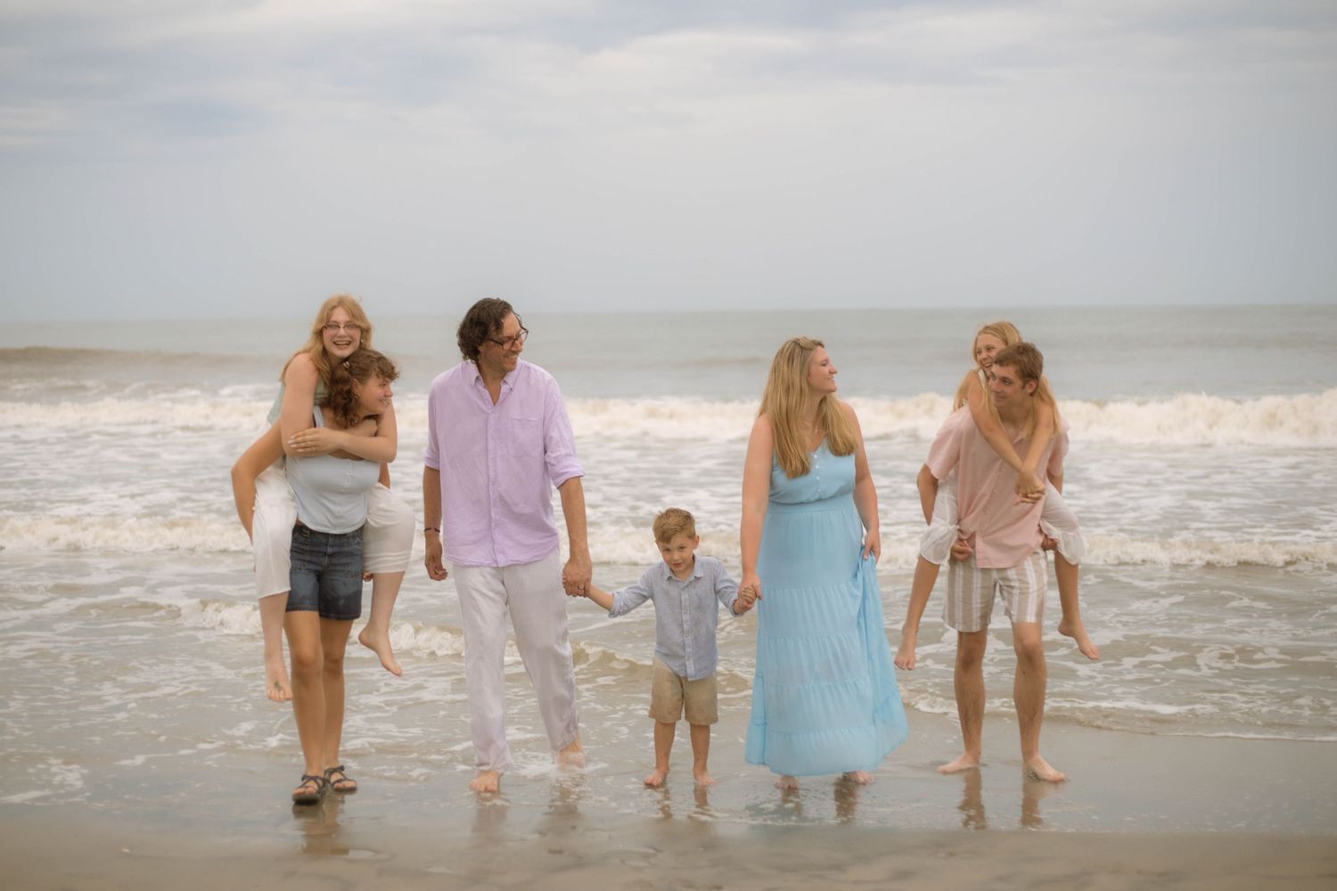 Family of six walking on a sandy beach near ocean waves, with adults carrying children on a cloudy day.