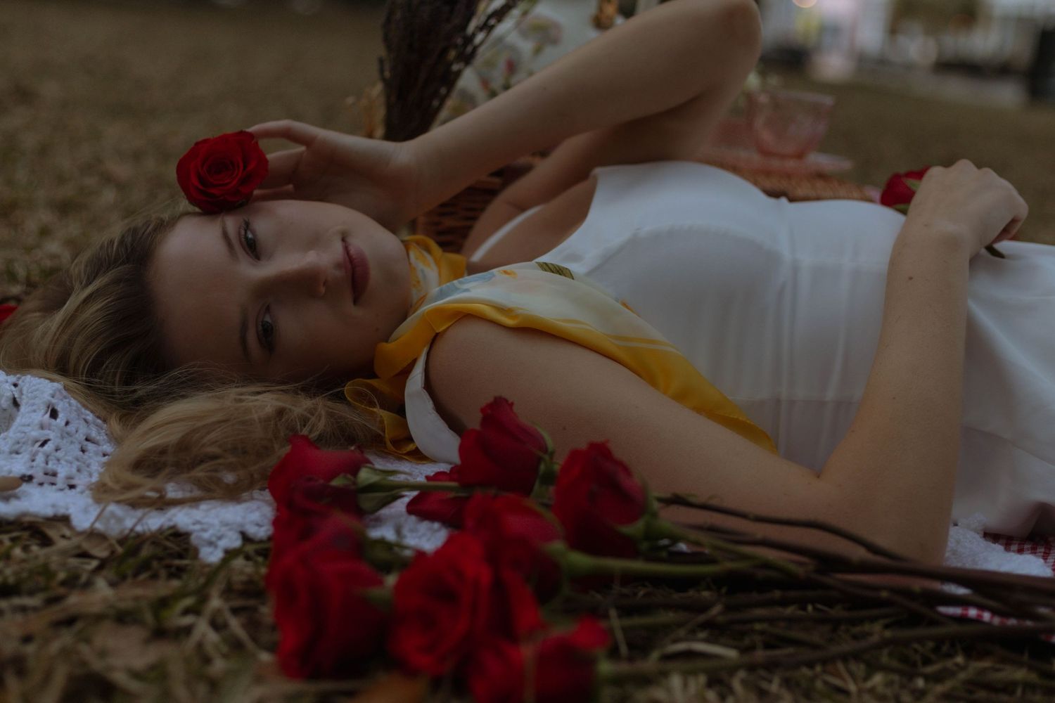 Red roses scattered beside a white dress on lace fabric creating a romantic outdoor setting.
