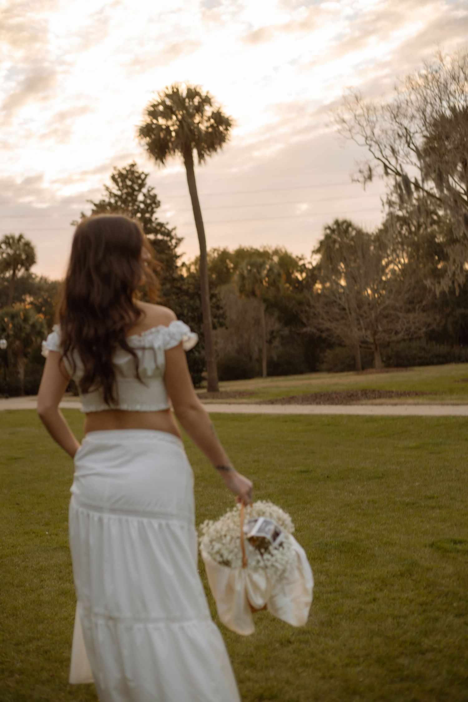Bride in white crop top and maxi skirt holds bouquet at sunset near palm trees on lush green lawn.