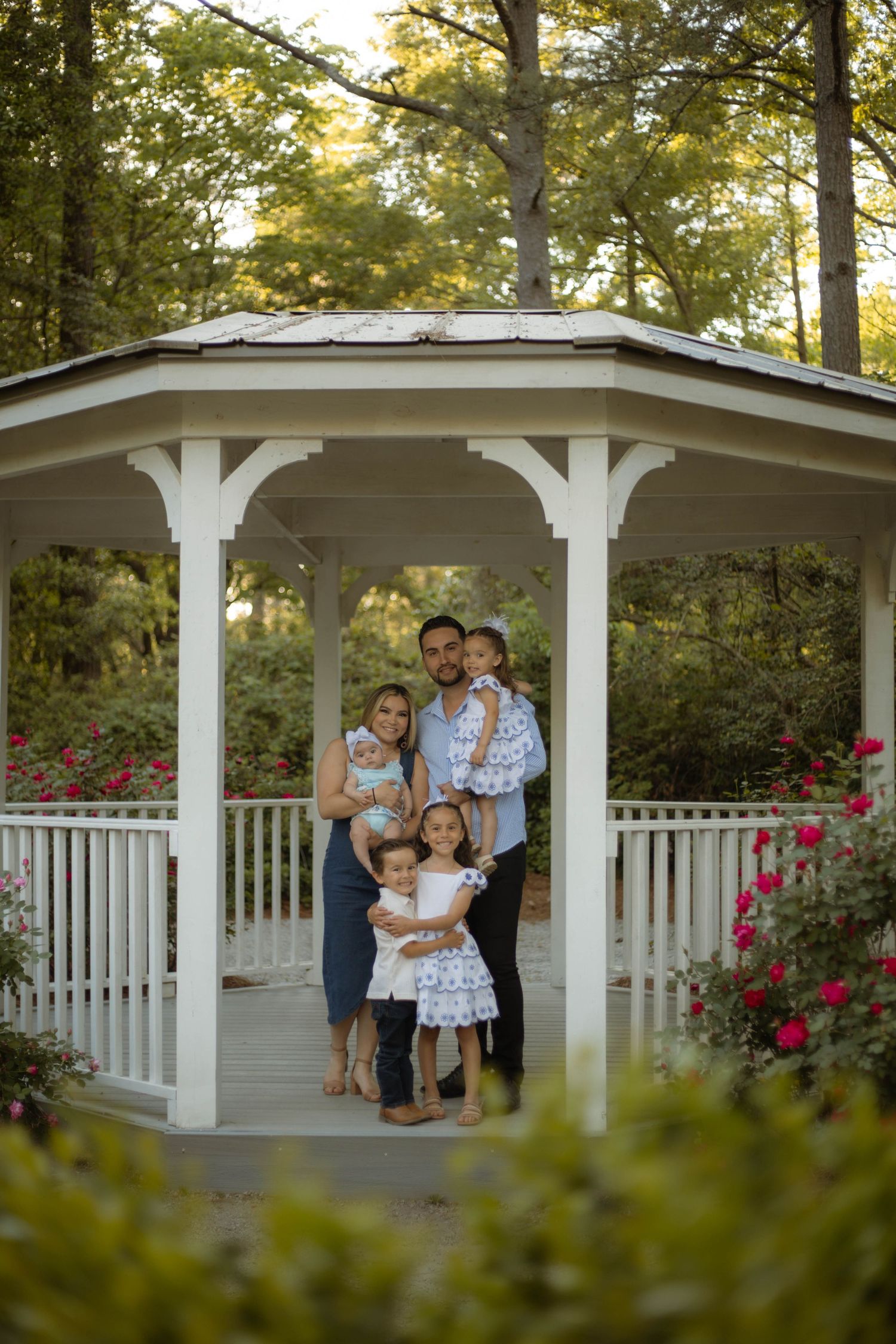 Family of five posing in a white gazebo surrounded by rose bushes and lush green trees during golden hour.