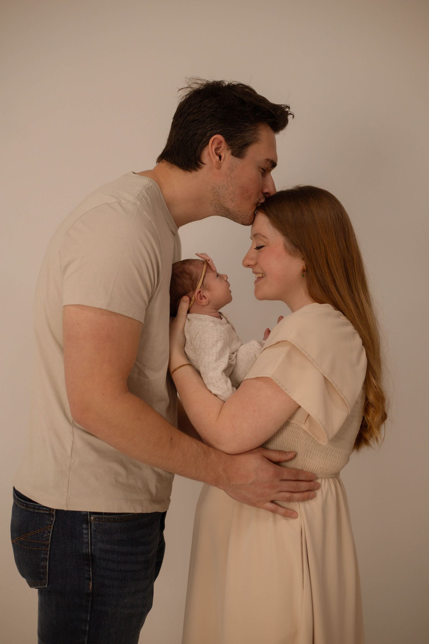 A young couple sharing a tender moment, kissing their newborn baby in a soft, neutral-toned studio portrait.