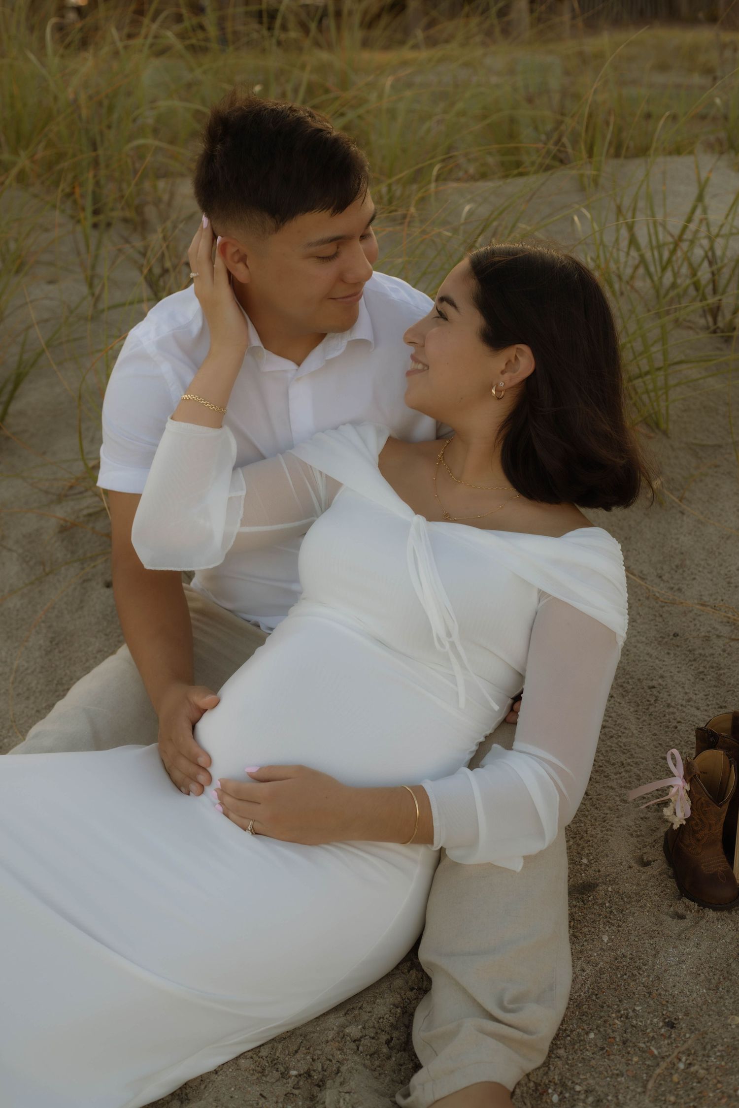 Pregnant couple in white outfits share a tender moment on sandy beach, woman cradling baby bump.