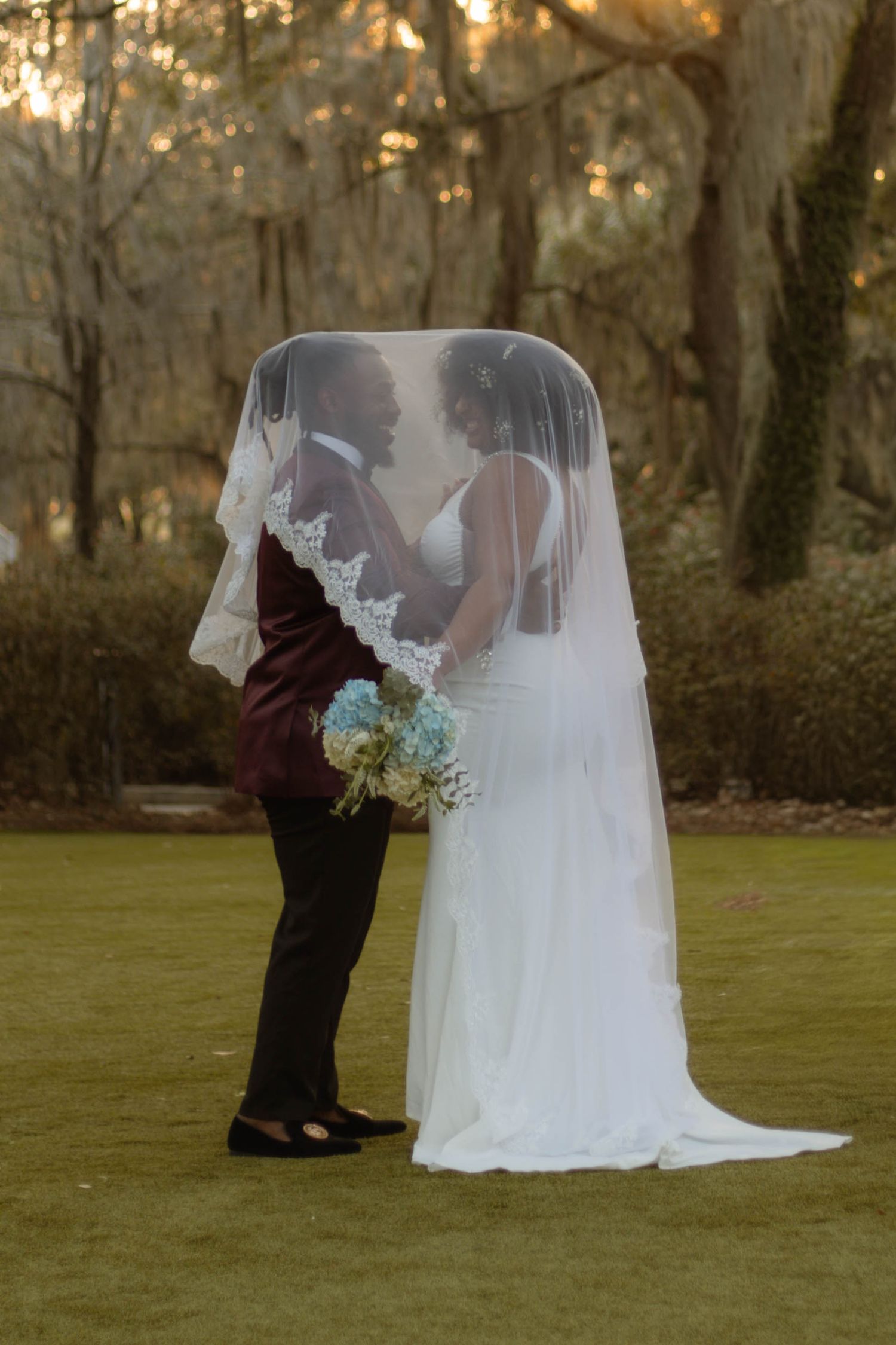 A couple in wedding attire share a romantic moment at sunset in a wooded outdoor setting.