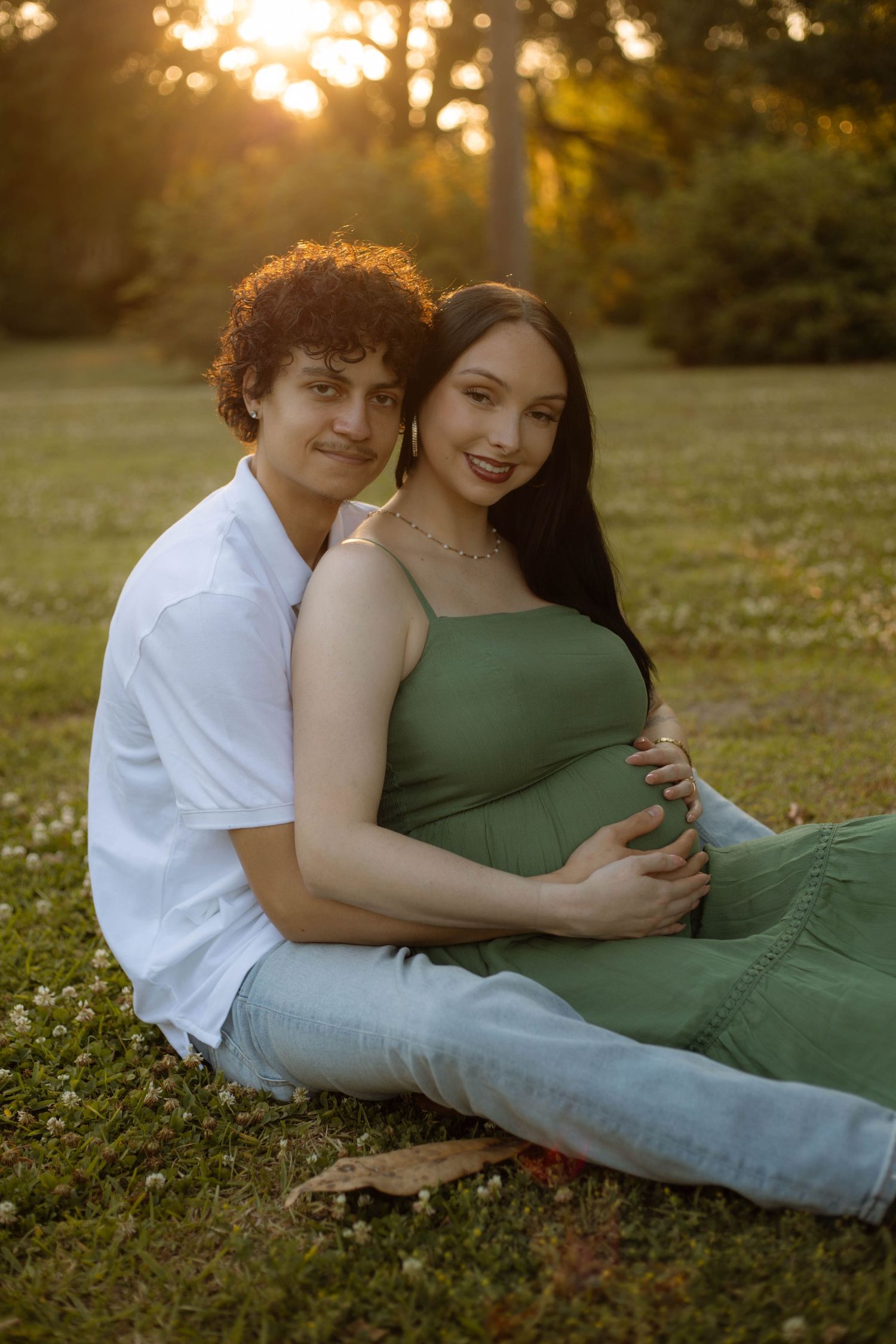 Couple sitting on grass at sunset, woman in green maternity dress cradling baby bump, man in white shirt embracing her.