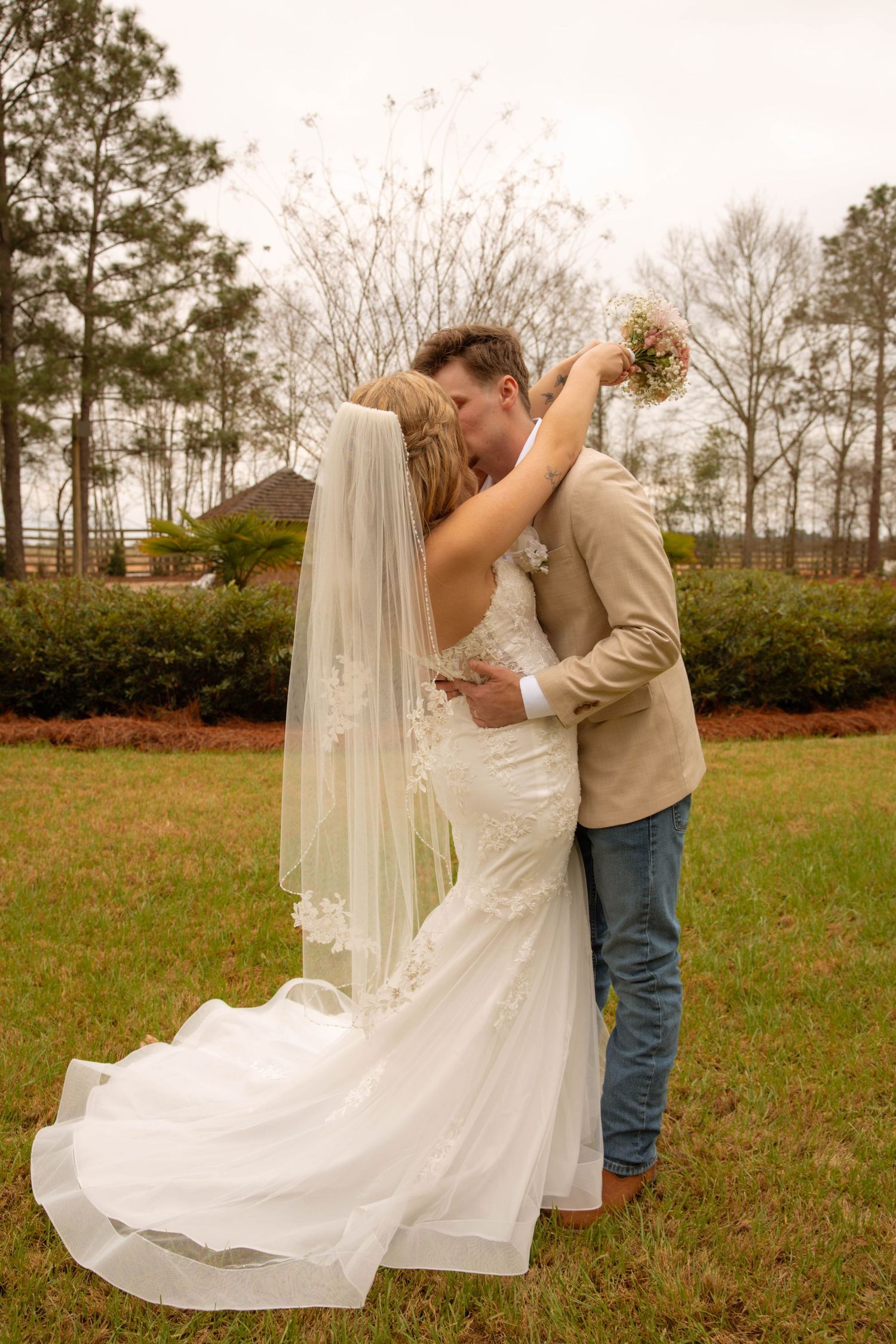 Bride and groom share a romantic kiss outdoors, bride in lace mermaid gown with veil, groom in tan blazer and jeans.