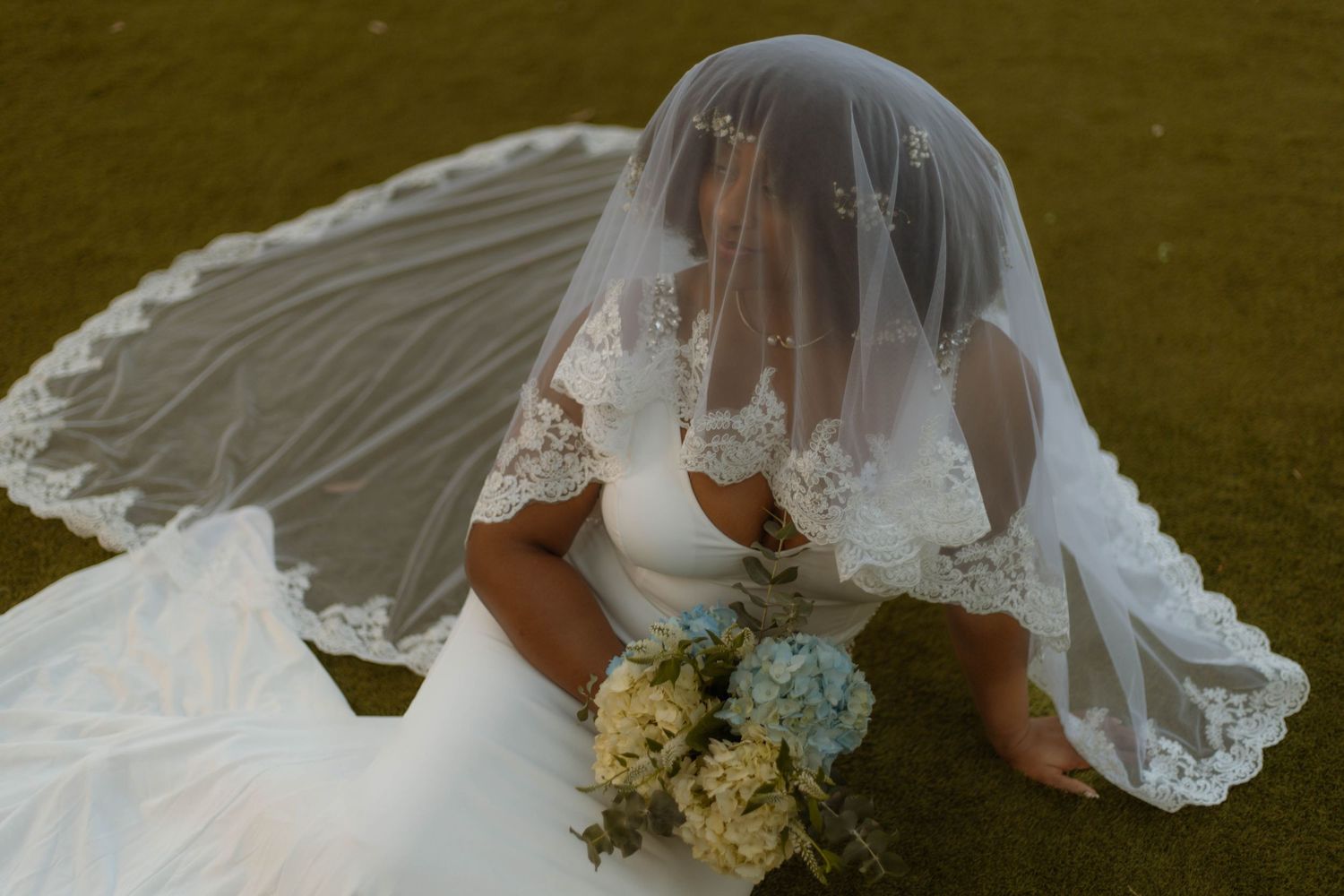 A bride in a flowing white wedding dress and veil holds a bouquet of cream and white flowers against a green background.