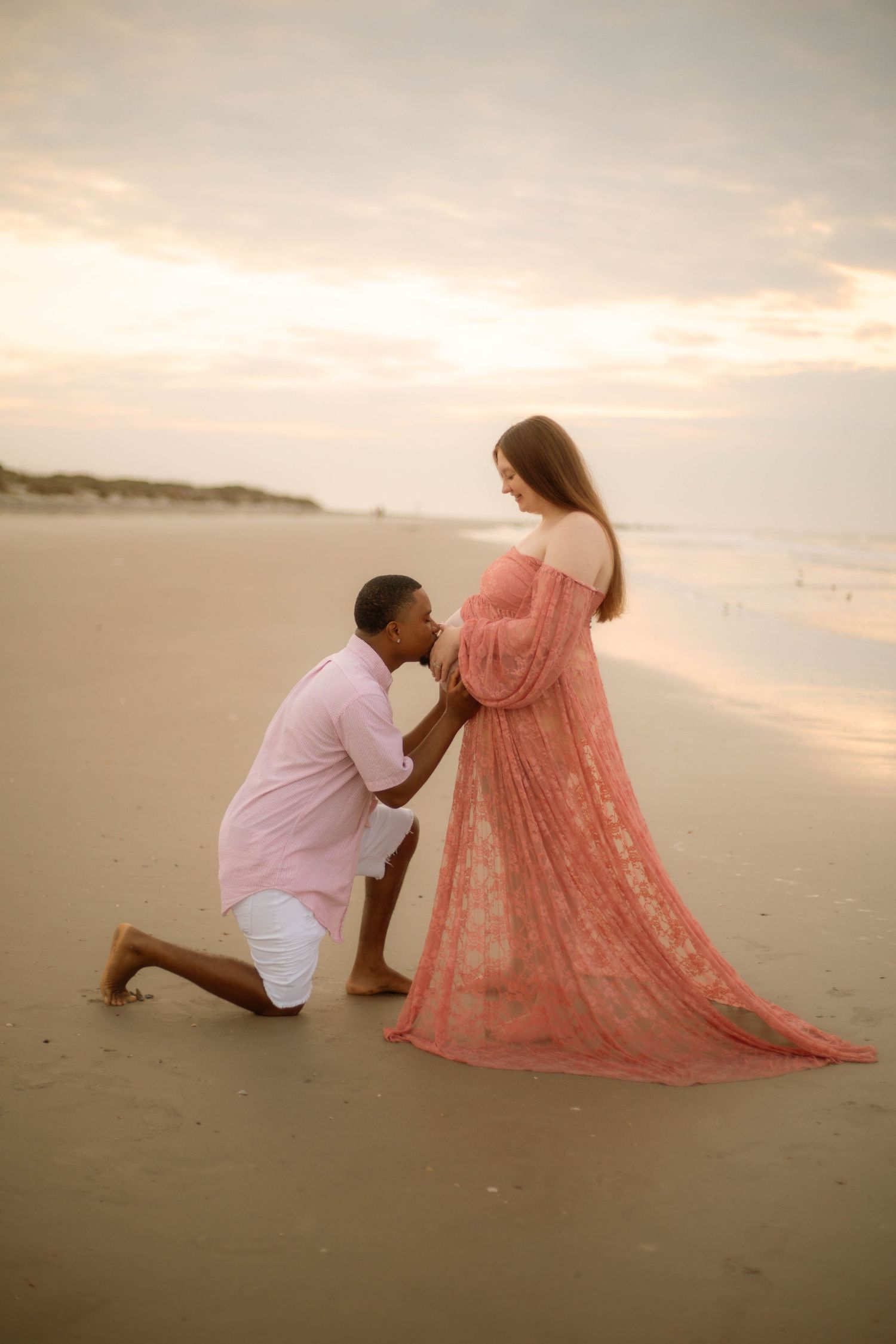 A man kneels on a beach before his pregnant partner in a flowing pink lace gown during a golden hour maternity photoshoot.