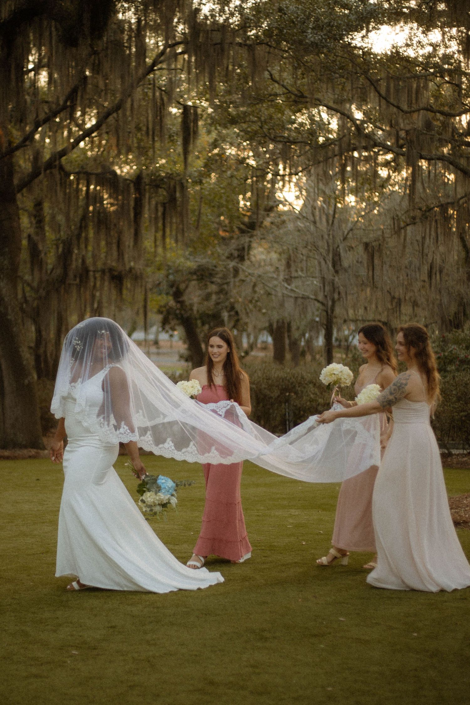 A wedding party in pastel dresses poses with a flowing veil in a sunlit garden with Spanish moss draped trees.