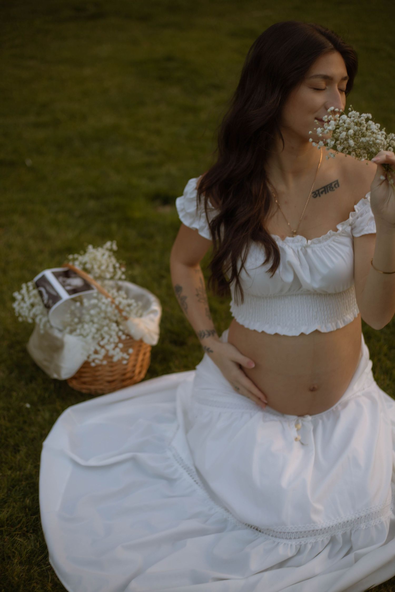 Pregnant woman in white crop top and skirt sitting on grass, smelling baby's breath flowers near a wicker basket.