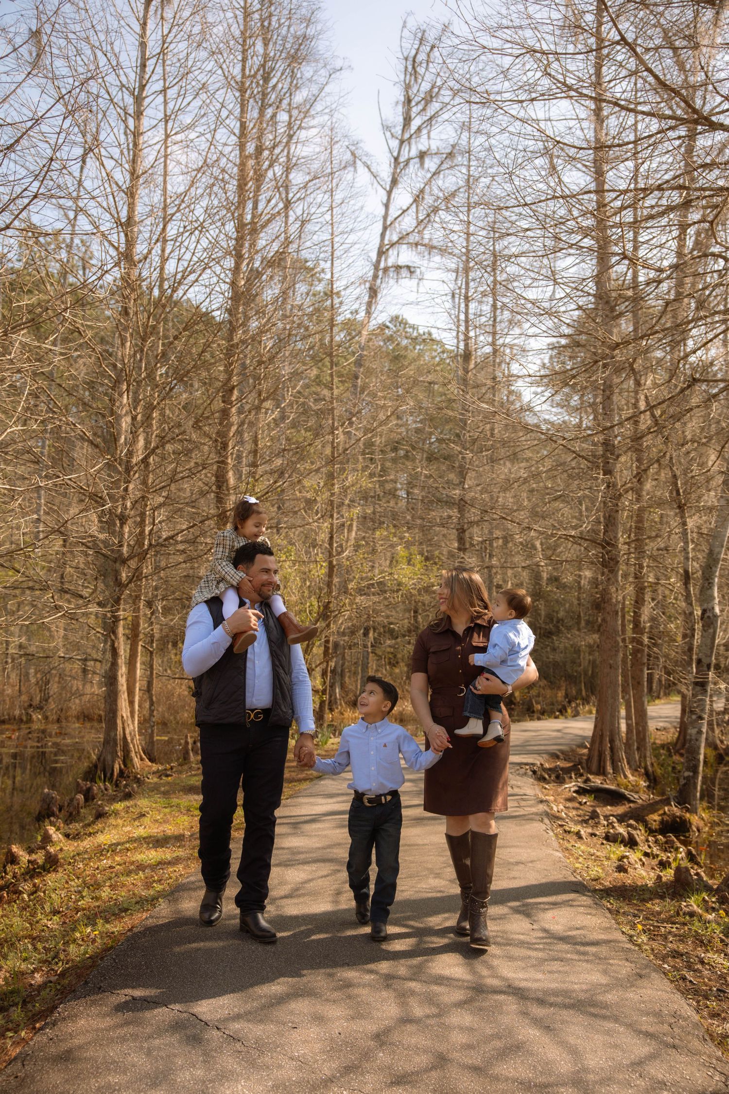 Family of five walking on a tree-lined path in autumn, parents carrying young children while middle child walks between them.
