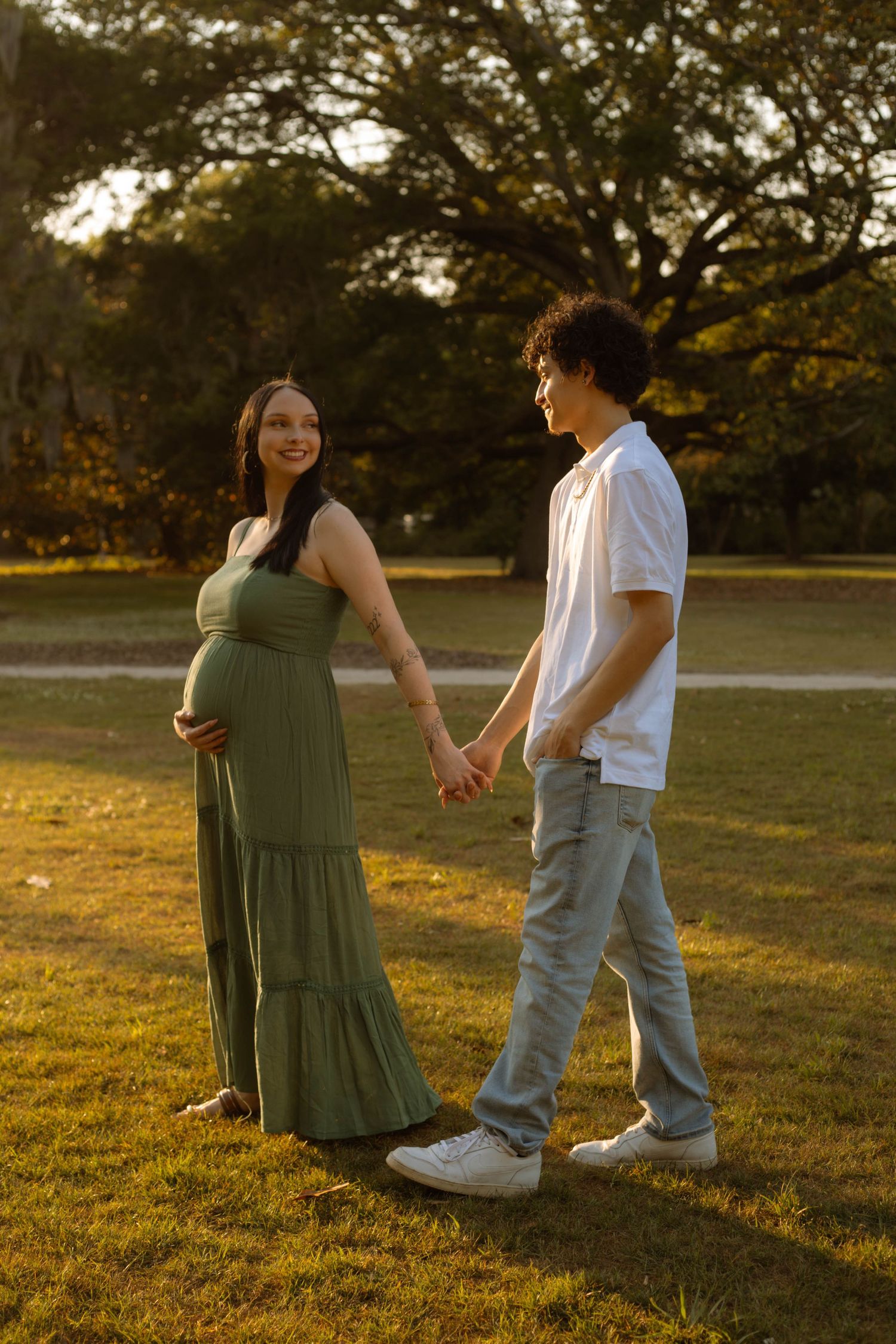 Pregnant couple holding hands in a sunlit park, woman in green maxi dress cradling baby bump, man in white shirt.