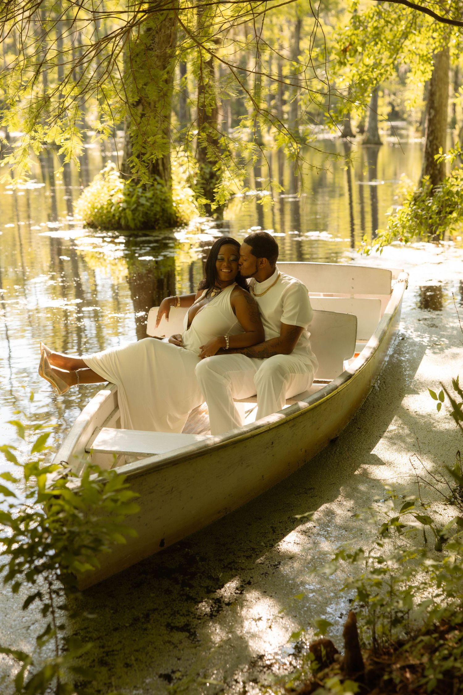 A pregnant couple in white outfits share a tender moment in a white rowboat surrounded by a serene, sunlit forest lake.
