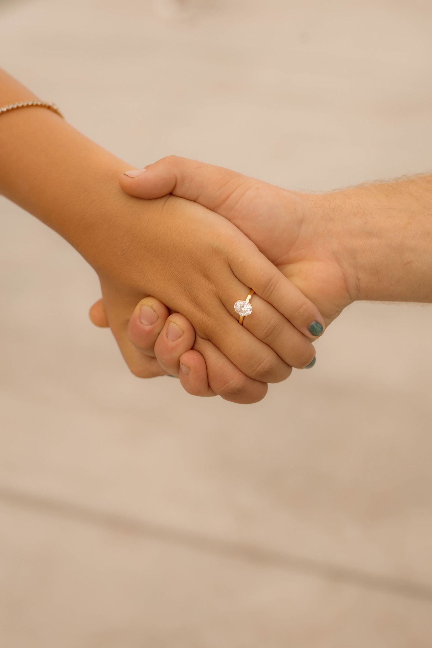 Two hands holding together, woman wearing a diamond engagement ring with teal nail polish and gold bracelet.