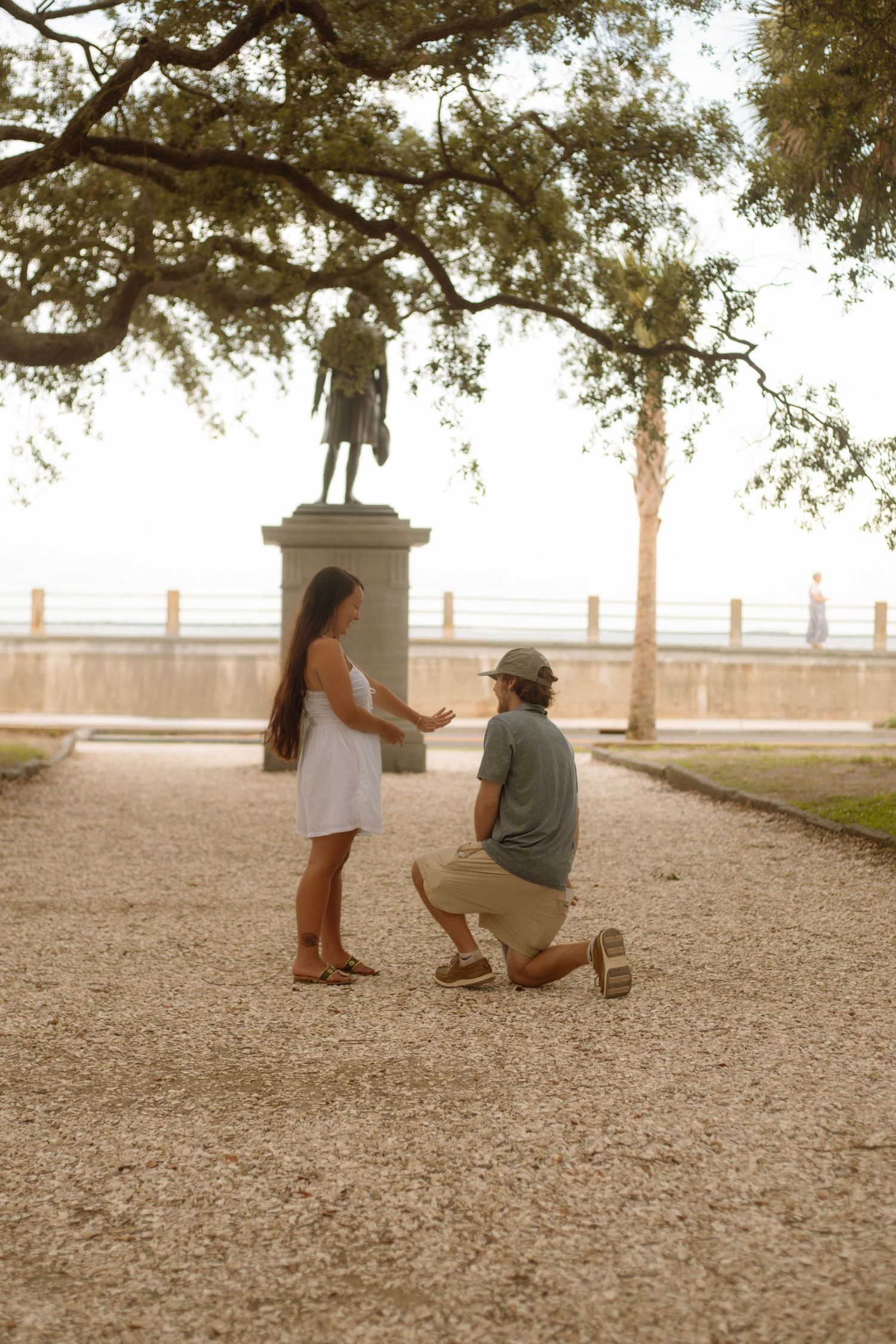 Man kneeling on one knee proposing to woman in white dress near historic statue at golden hour.