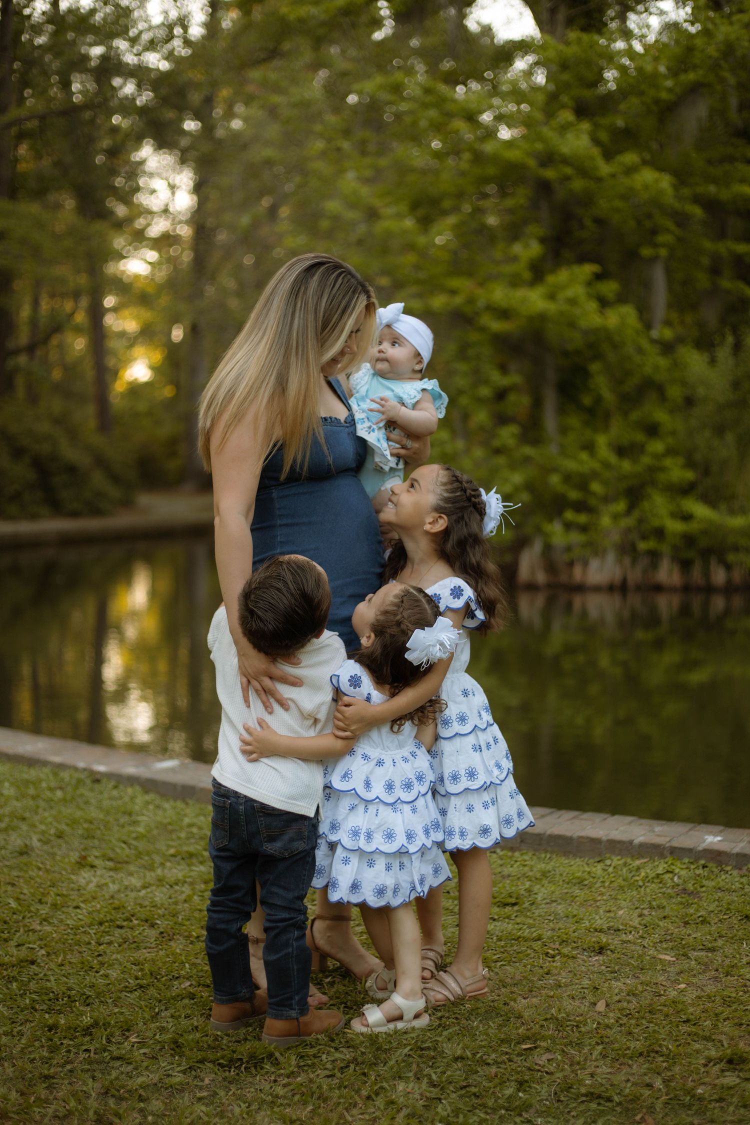 Pregnant woman holding baby surrounded by three young children hugging her near a serene pond in a lush green park.