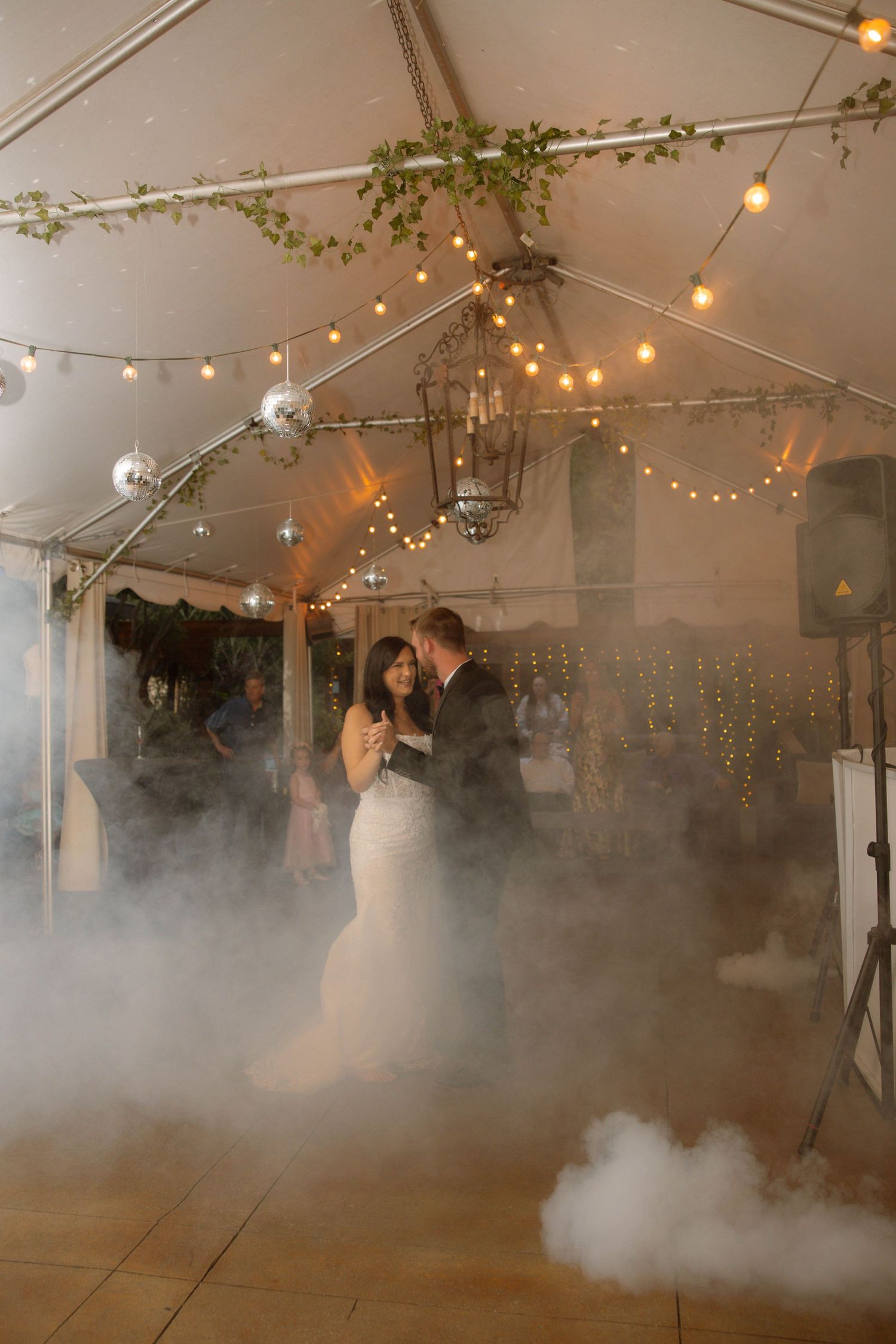 Bride and groom share first dance surrounded by dramatic fog under string lights in a tent venue.