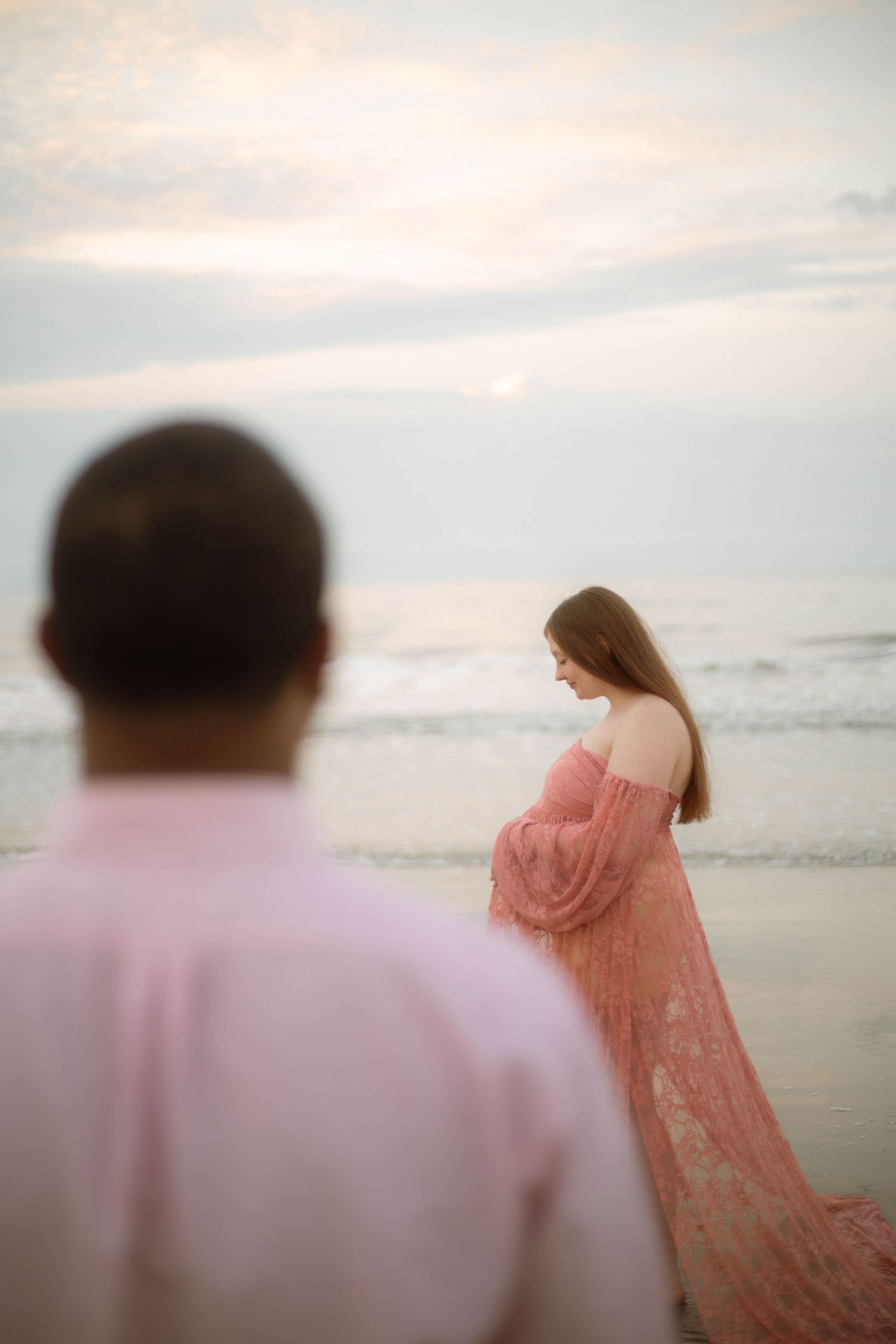 Pregnant woman in pink lace gown stands at beach during sunset while man in pink shirt watches from foreground.