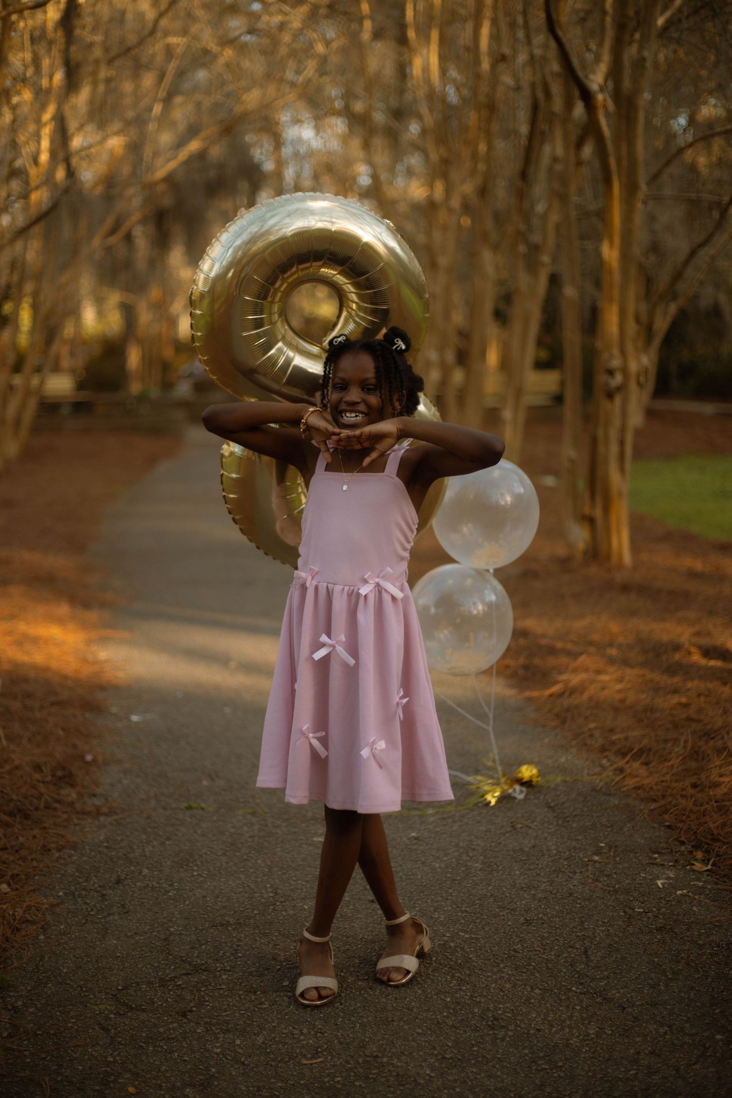A young girl in a pink dress holds gold and white balloons on a tree-lined path, smiling joyfully.