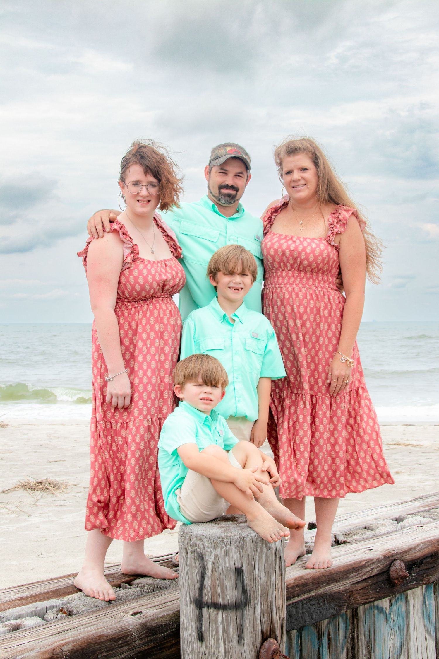 A family wearing coordinated coral and mint outfits poses playfully on a wooden pier at the beach.