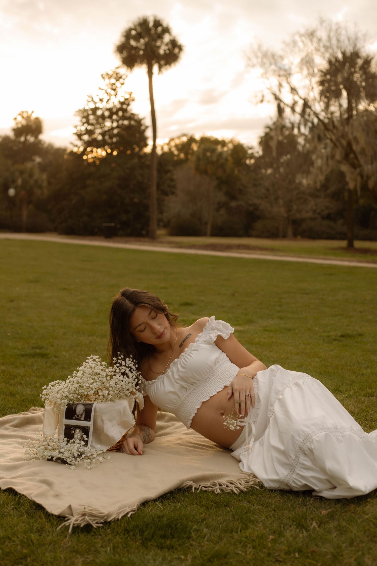 Pregnant woman in white outfit reclines on blanket in park at sunset with ultrasound photos and flowers.