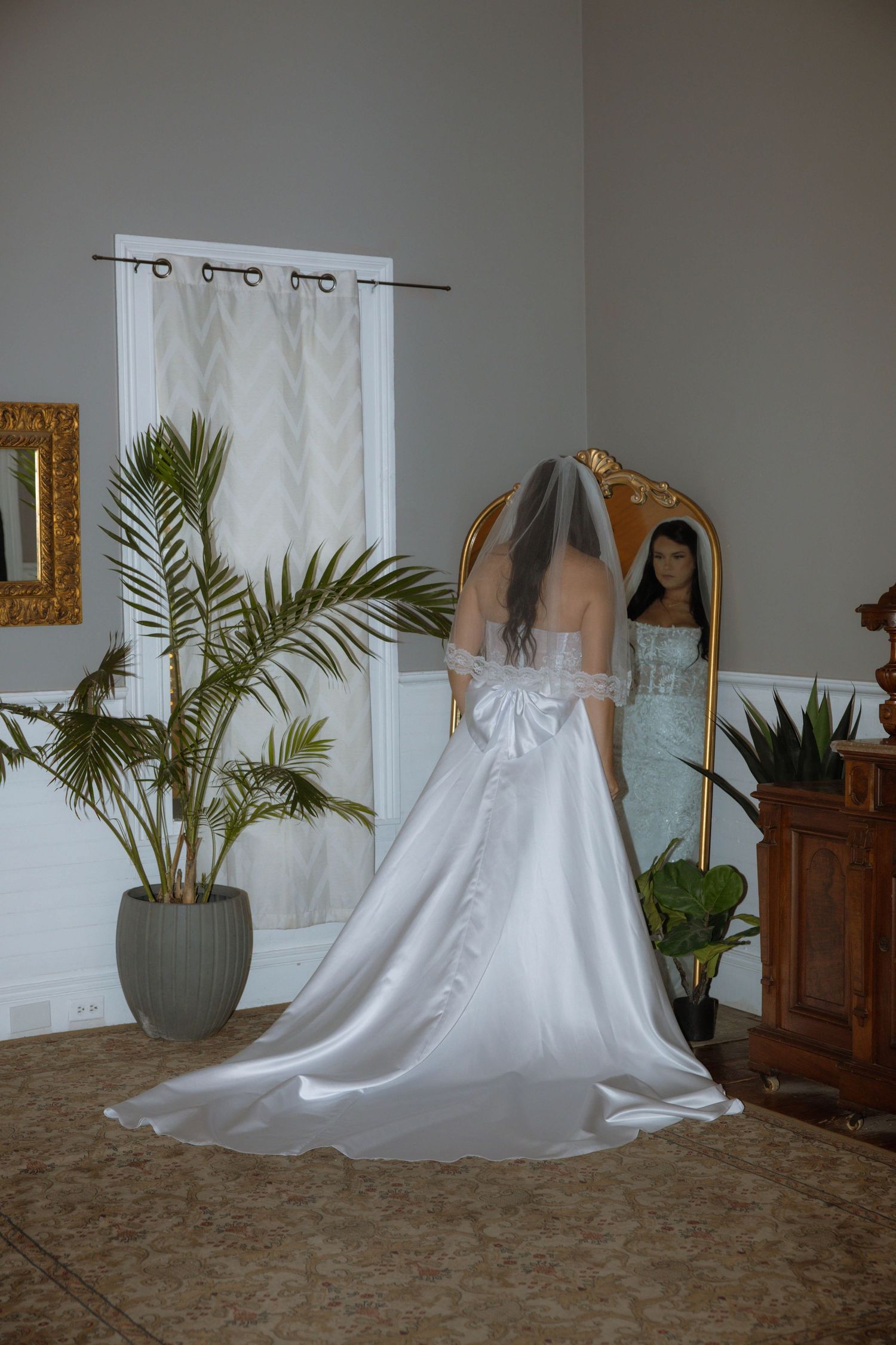 Bride in white wedding gown with veil and bow back detail stands before ornate gold mirror in elegant room.