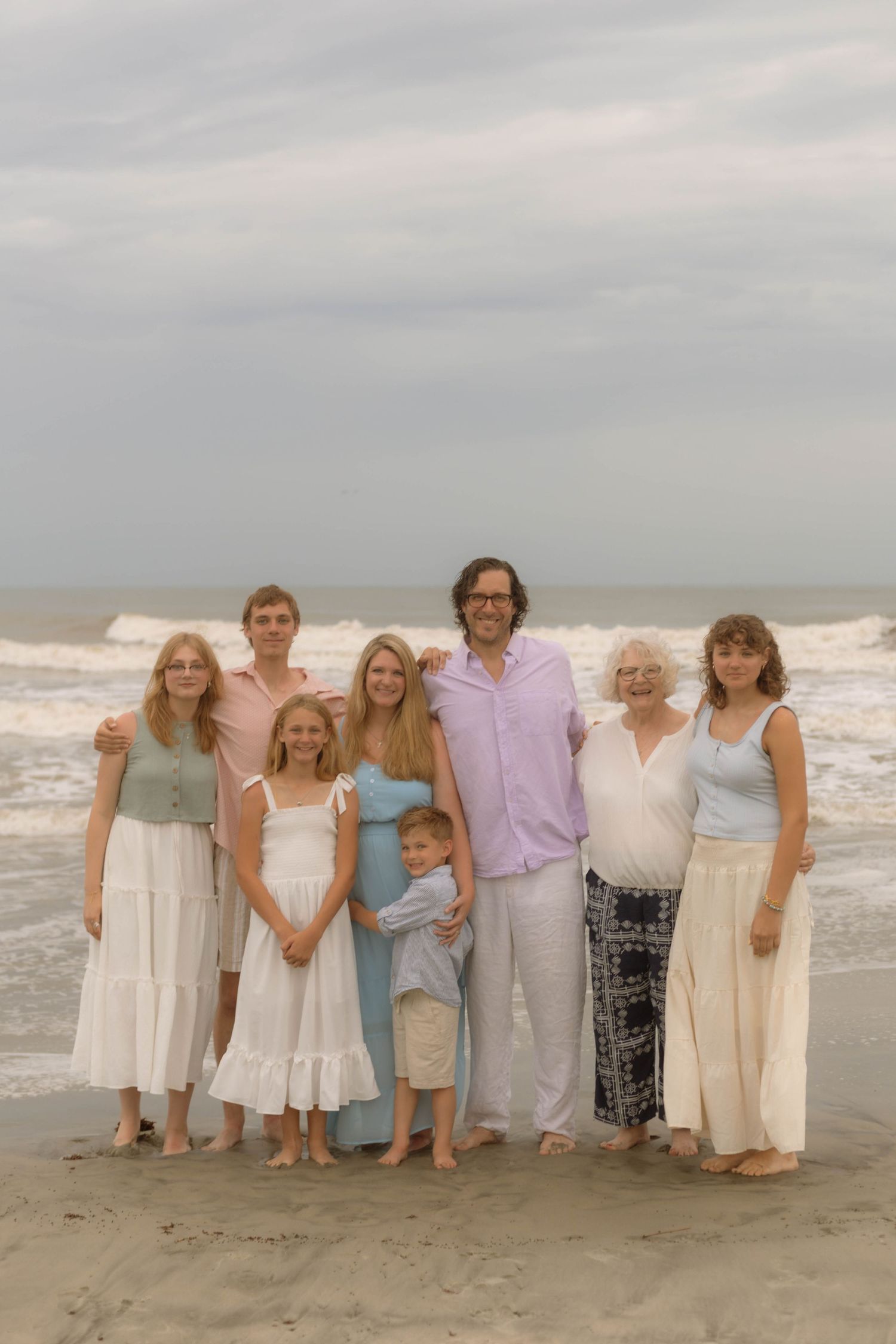 Multi-generational family of eight poses together on a sandy beach with ocean waves behind them on a cloudy day.