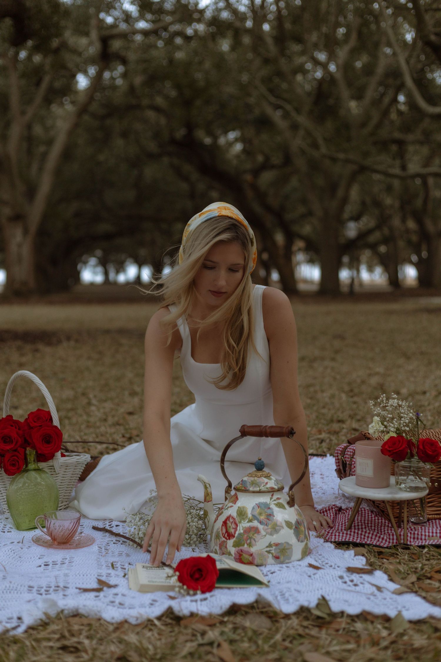 A picturesque outdoor picnic scene with white linens, red roses and vintage decor arranged on grassy ground beneath trees.