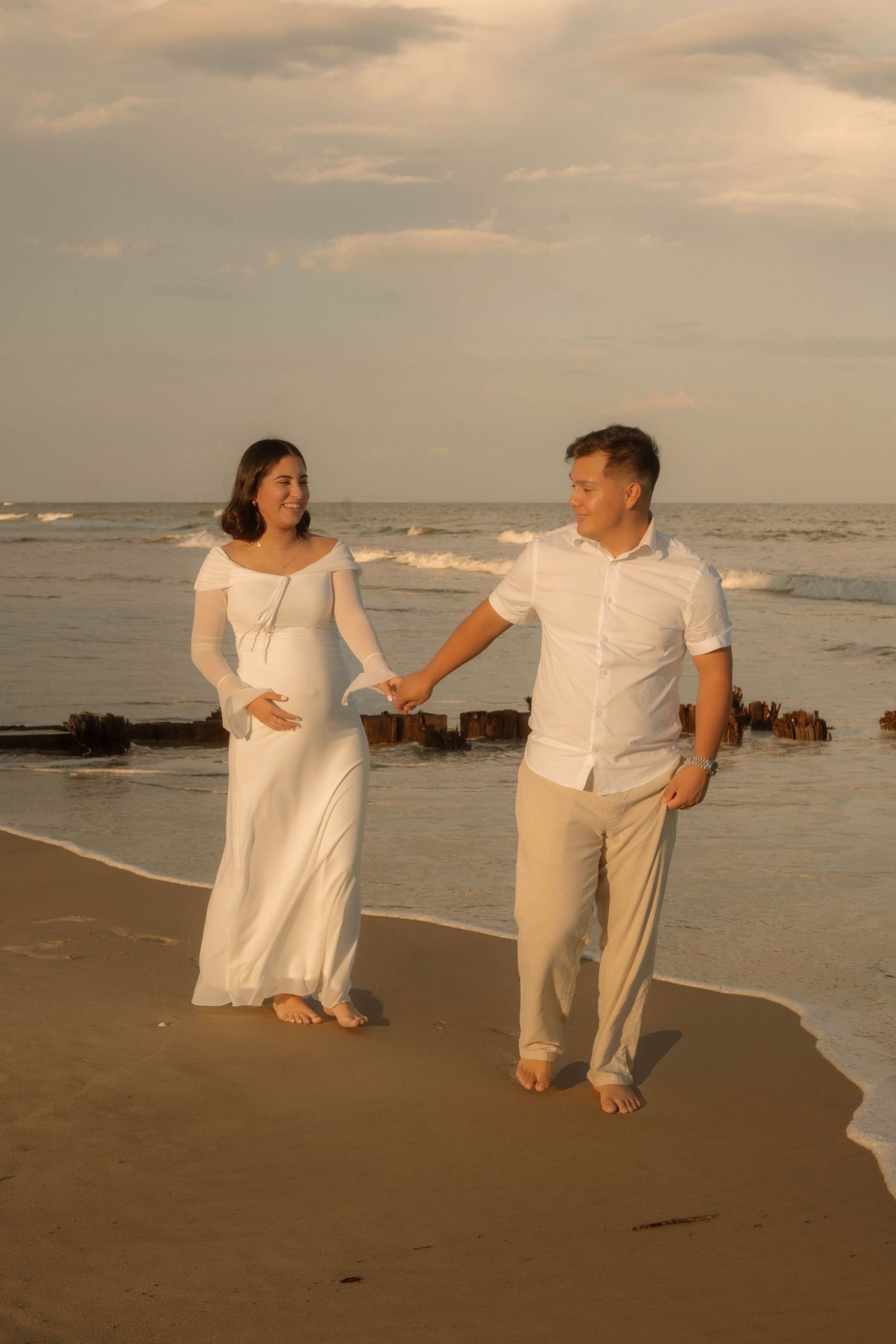Pregnant couple holding hands on beach at sunset, woman in white dress, man in white shirt and beige pants.